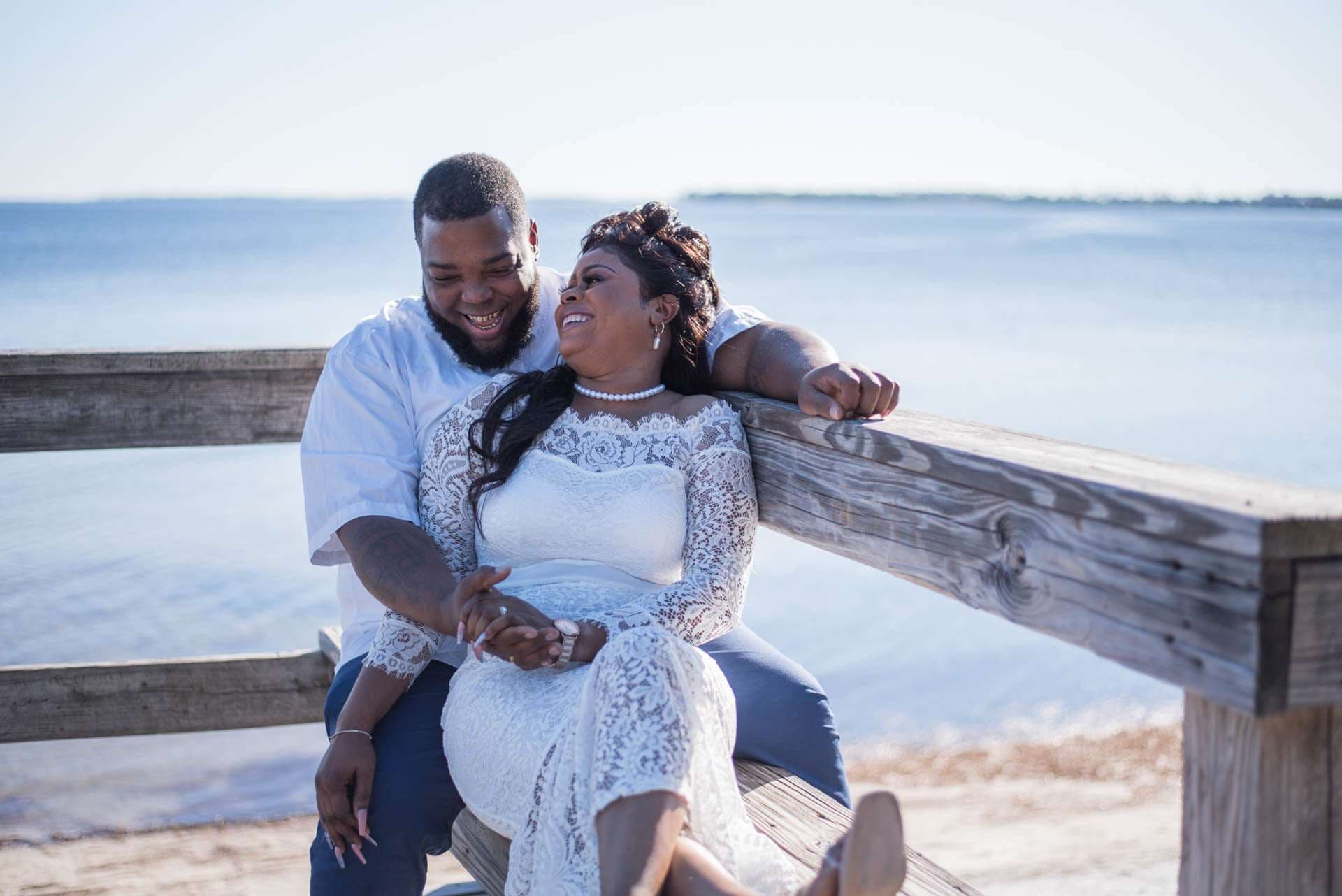 Couple laughing together on a wooden bench by the bay, bride in a white lace dress leaning against her groom