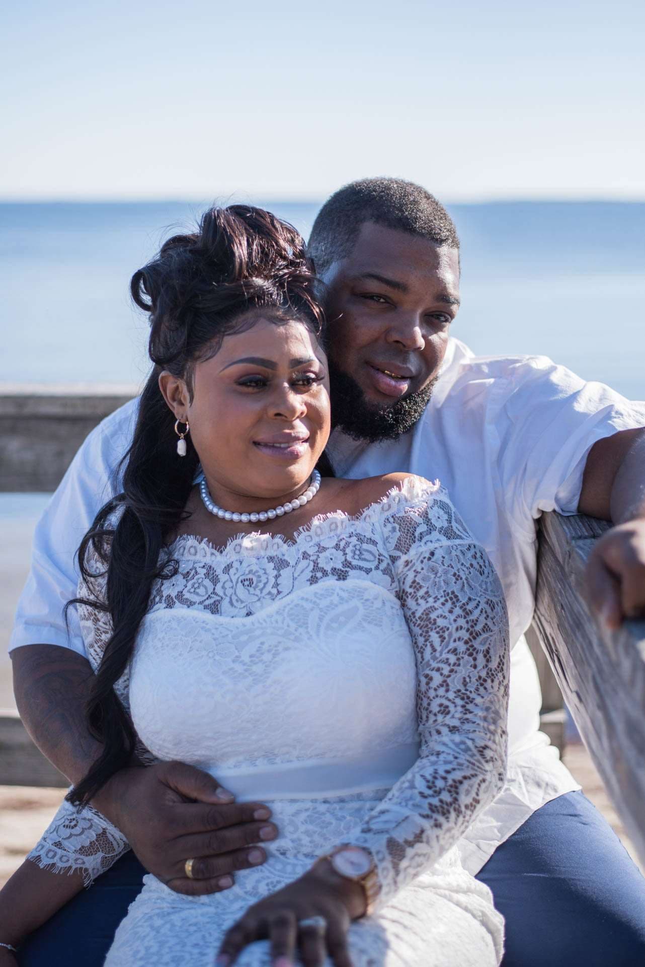 Couple embracing on a wooden bench by the water, woman in a white lace off-shoulder dress and pearl necklace