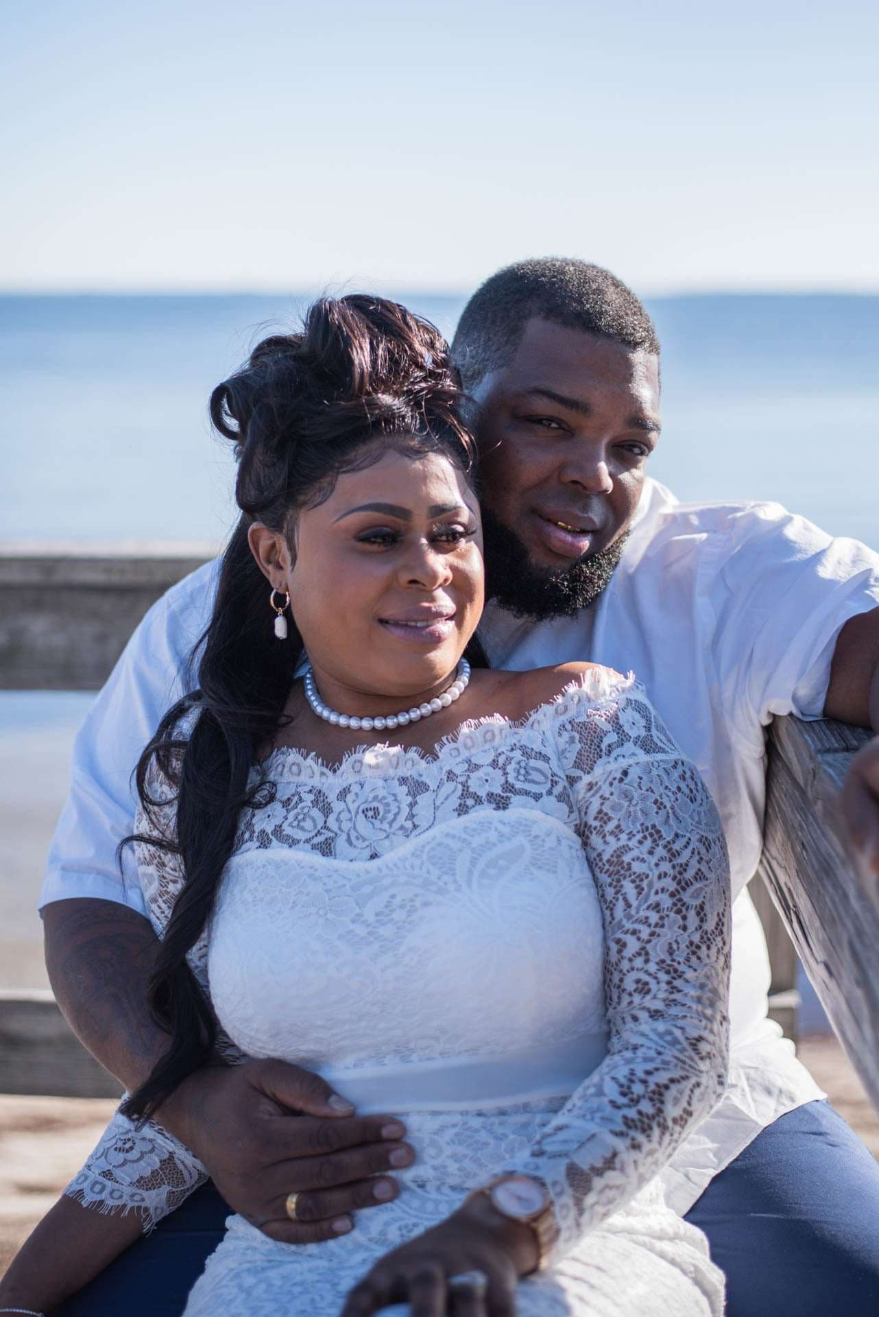 Couple embracing on a wooden bench by the water, woman in a white lace gown and pearl necklace