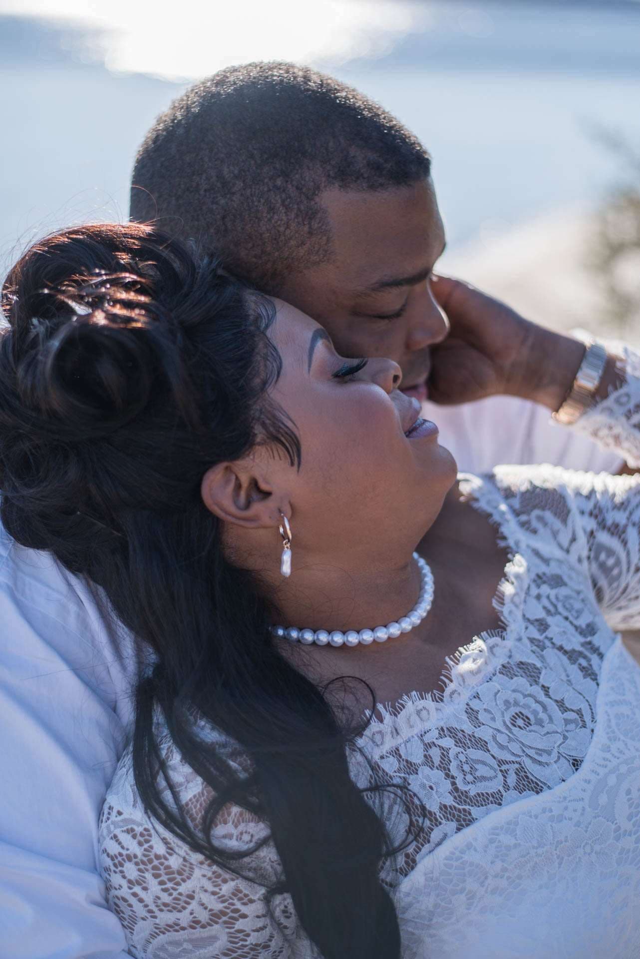 Couple embracing at a waterside elopement, the bride in a white lace dress and pearl necklace with her eyes closed
