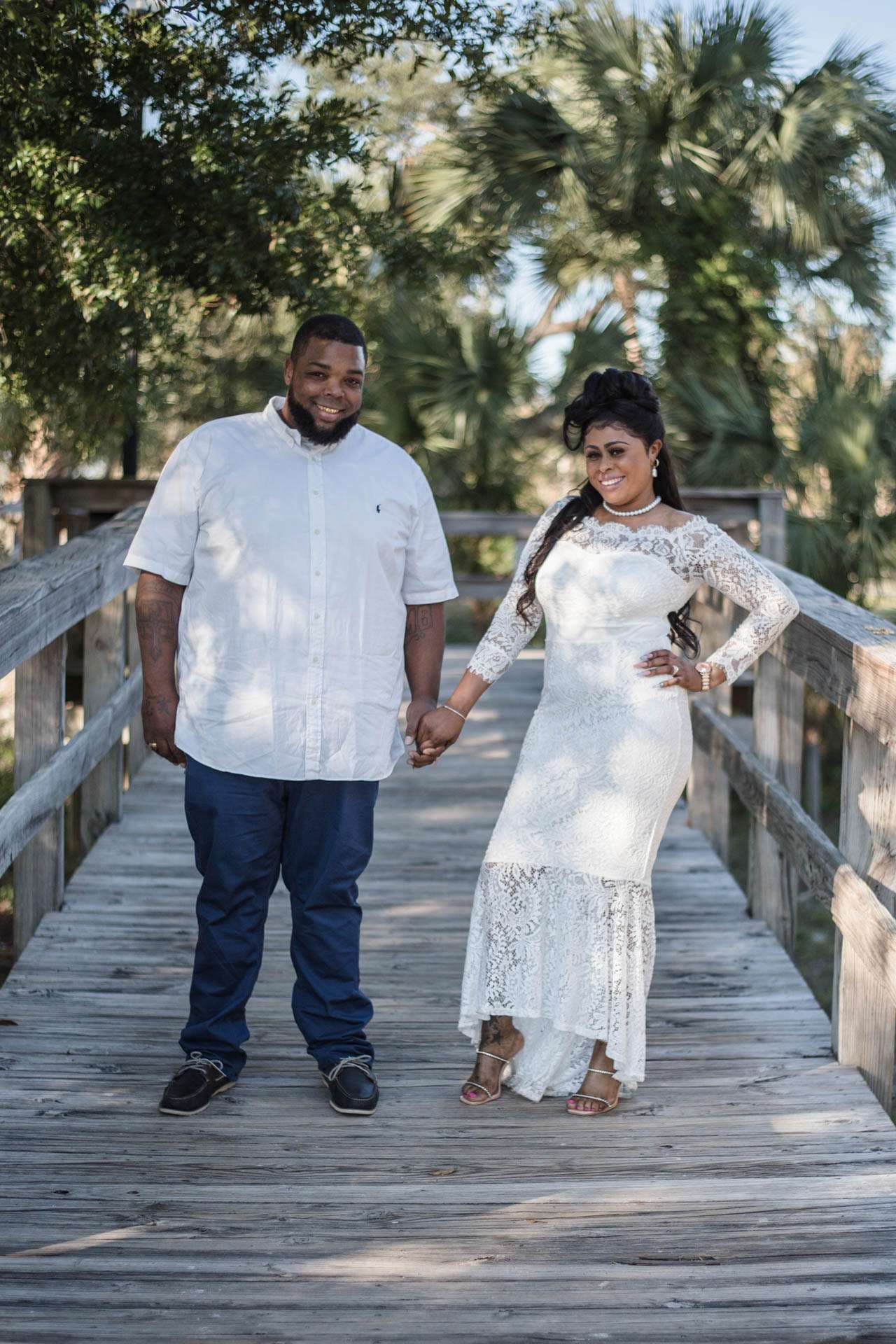 Couple holding hands on a wooden boardwalk among palm trees, he in a white shirt, she in a white lace gown