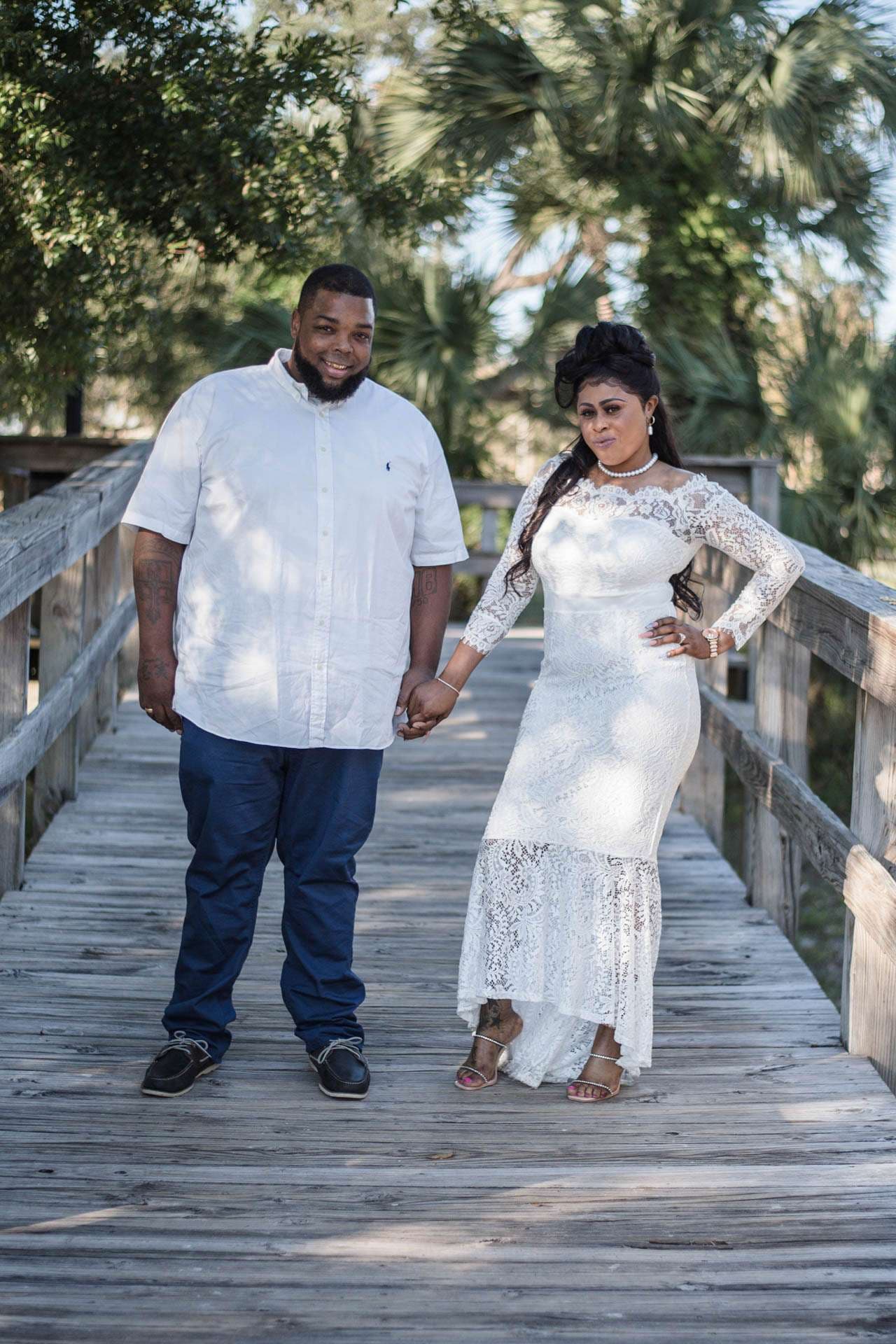 Couple holding hands on a wooden boardwalk among palm trees, he in a white shirt and jeans, she in a white lace gown