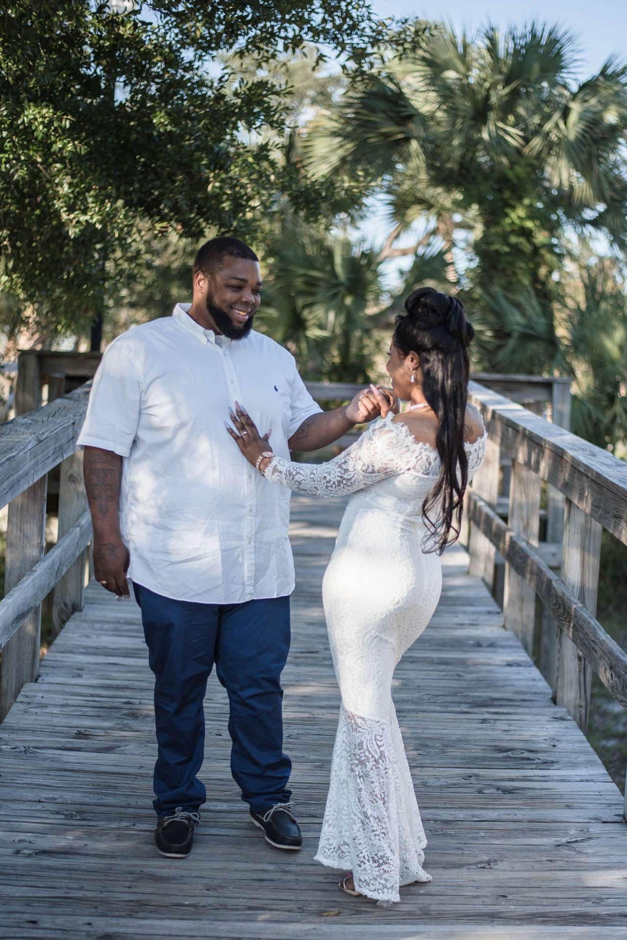 Smiling couple dancing on a wooden boardwalk, she in an off-shoulder white lace gown, he in white shirt and navy pants