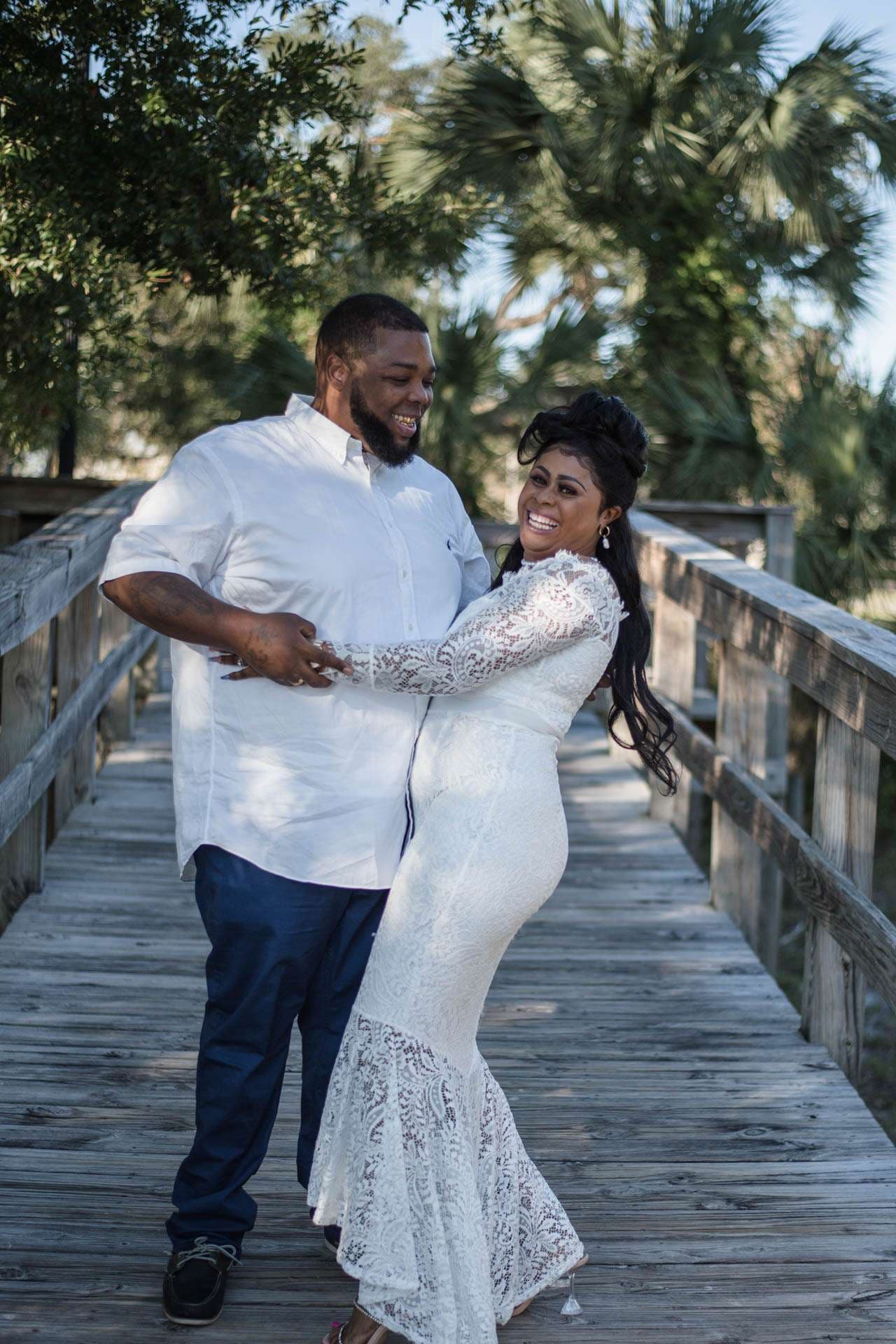 Smiling couple embracing on a wooden boardwalk, she in a white lace gown, he in a white shirt and blue pants