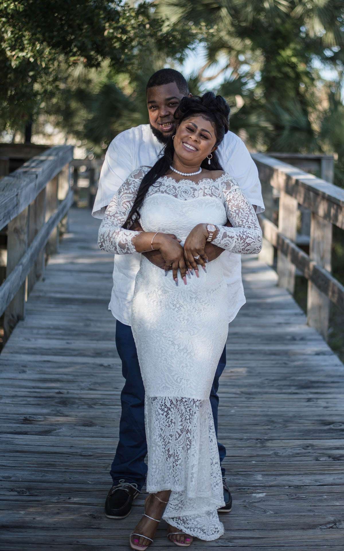 Man hugging a smiling woman in a white lace gown from behind on a wooden boardwalk among trees