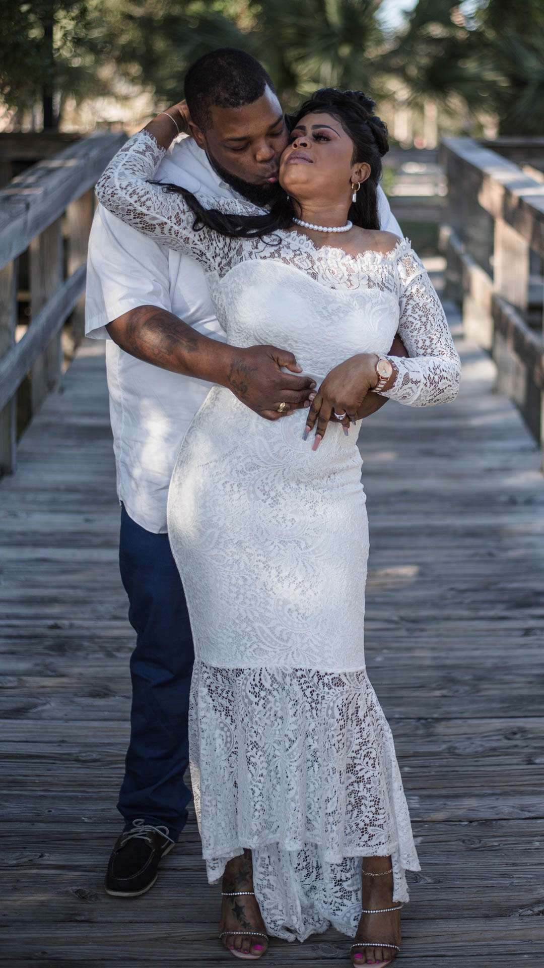 Man kissing a woman's forehead as he embraces her from behind on a wooden boardwalk, she wears a white lace gown