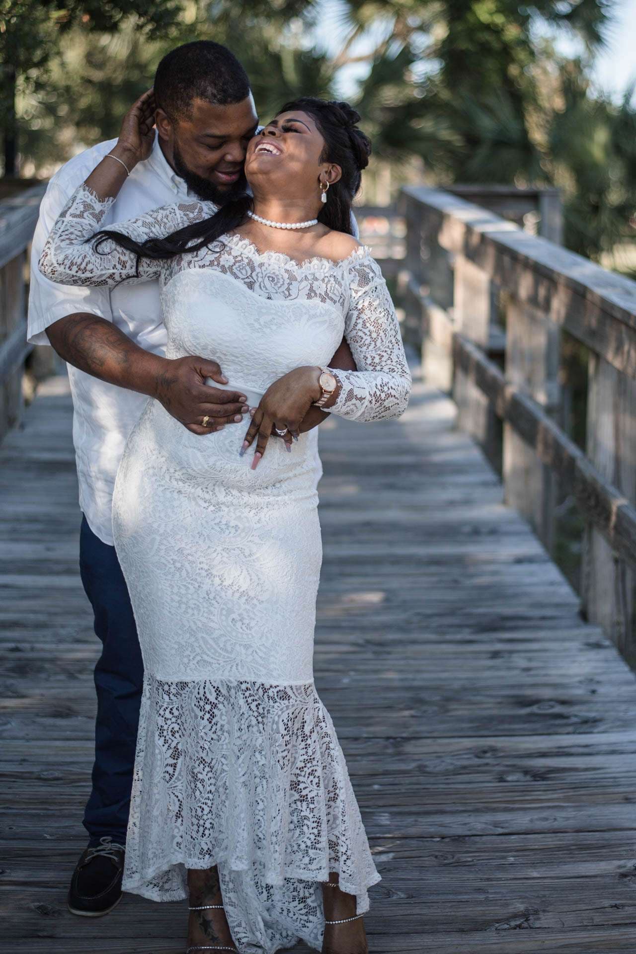 Groom hugging his laughing bride from behind on a wooden boardwalk, bride in a white lace gown
