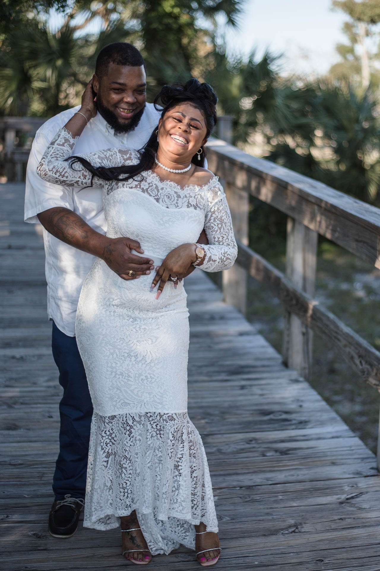 Laughing bride in a white lace gown embraced from behind by her groom on a weathered wooden boardwalk