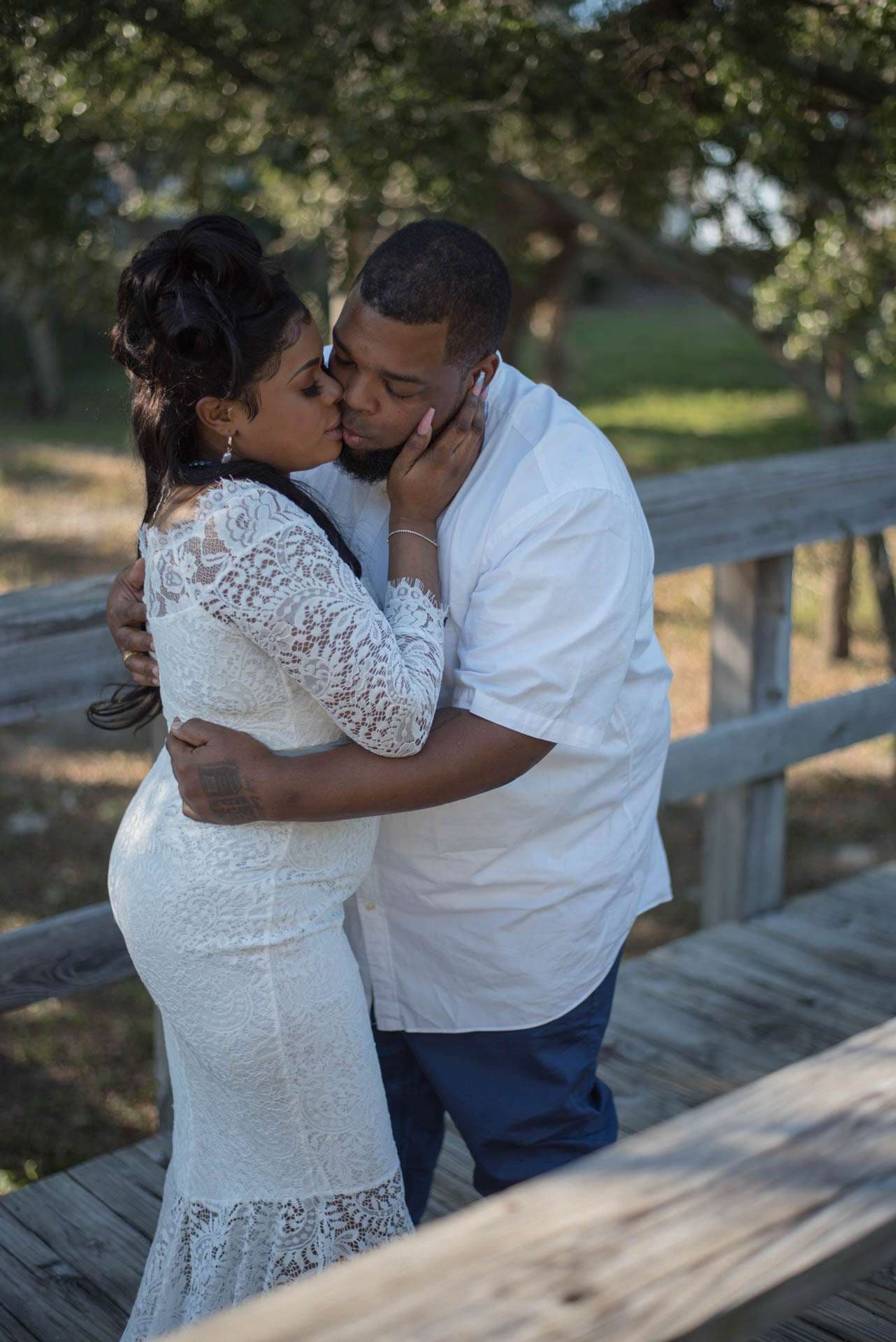 Couple embracing and kissing on a wooden boardwalk under trees at Oaks By The Bay, she in a white lace dress