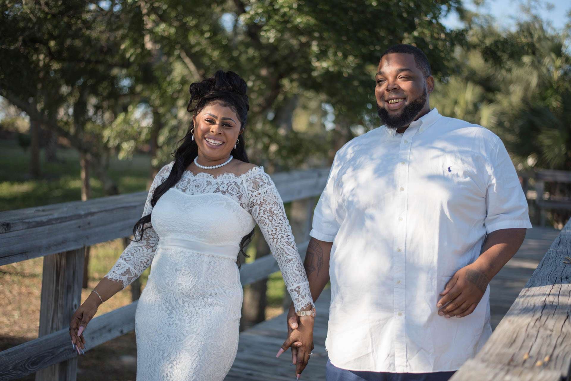 Smiling couple holding hands on a wooden boardwalk, she in a white lace dress and pearls, he in a white shirt