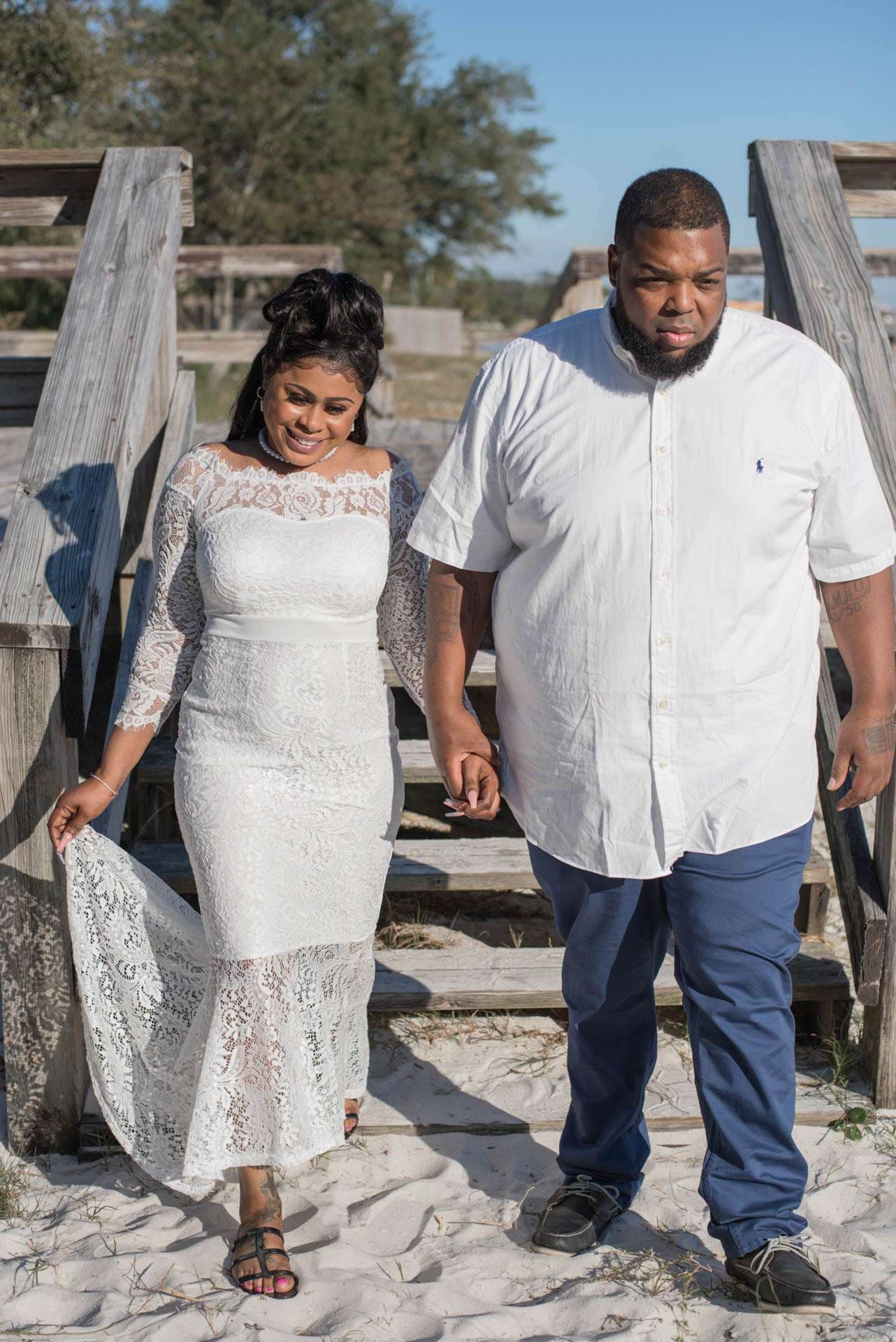 Smiling bride in a white lace gown holding her skirt walks hand in hand with her partner down weathered wooden beach steps
