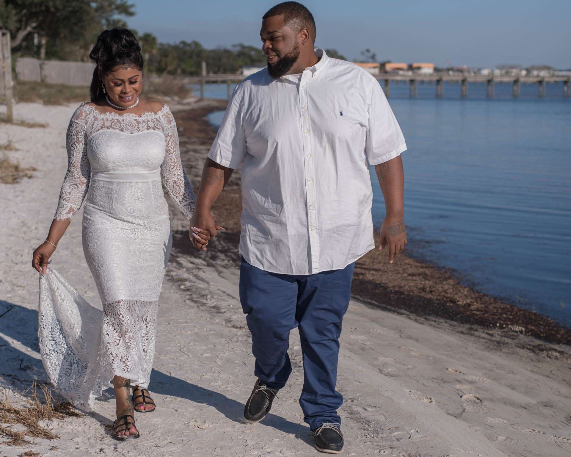 Couple holding hands and walking along a sandy shoreline, the bride in a white lace gown, groom in white shirt and blue pants