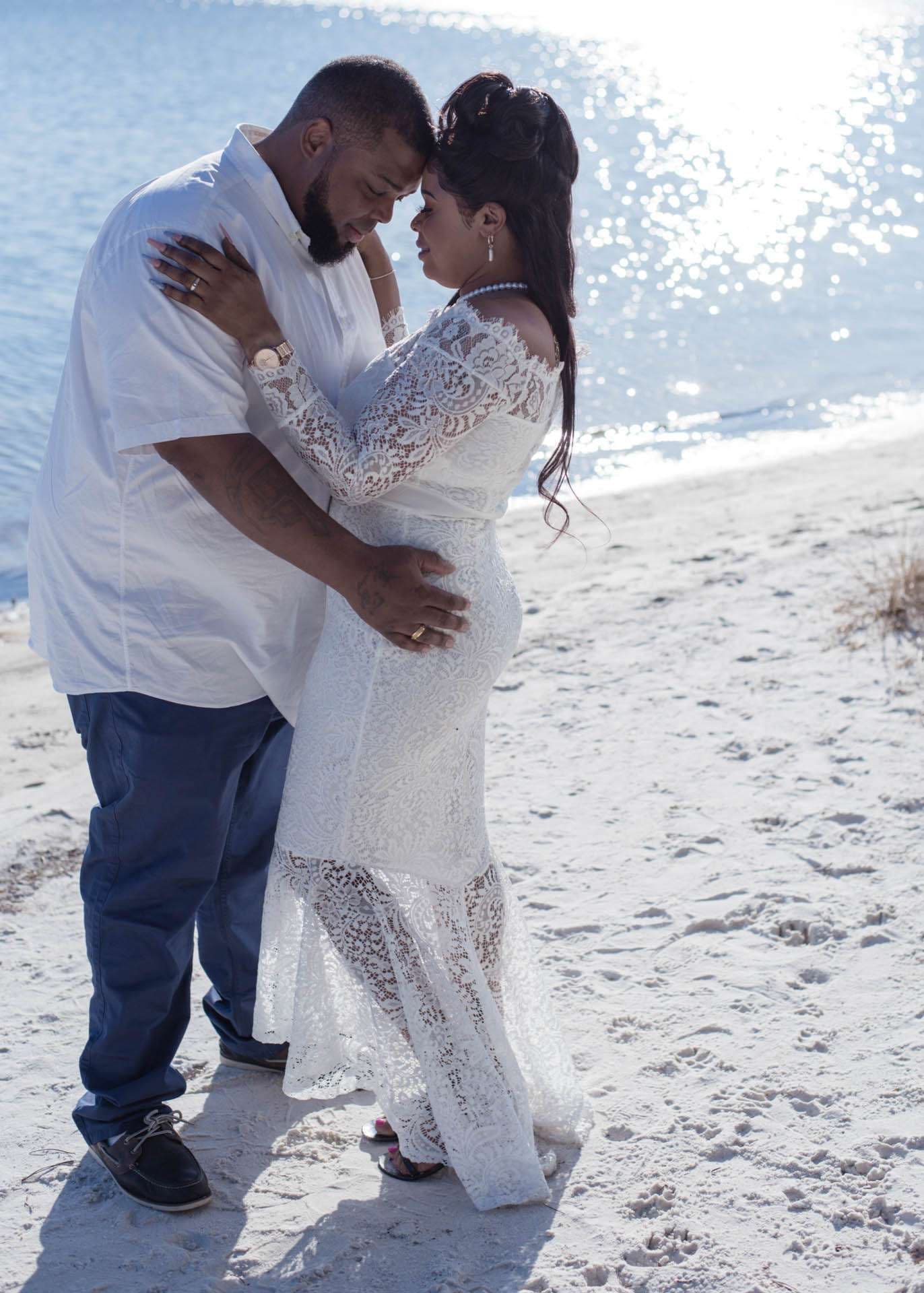 Couple embracing forehead to forehead on a sunny sandy beach, she in a white lace gown, he in a white shirt
