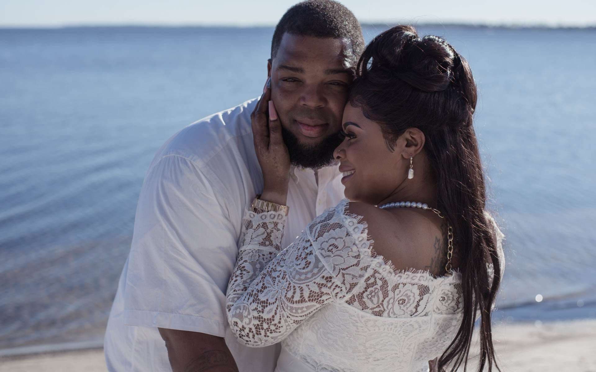 Couple embracing by the water, she in a white lace gown touching his cheek, he in a white shirt