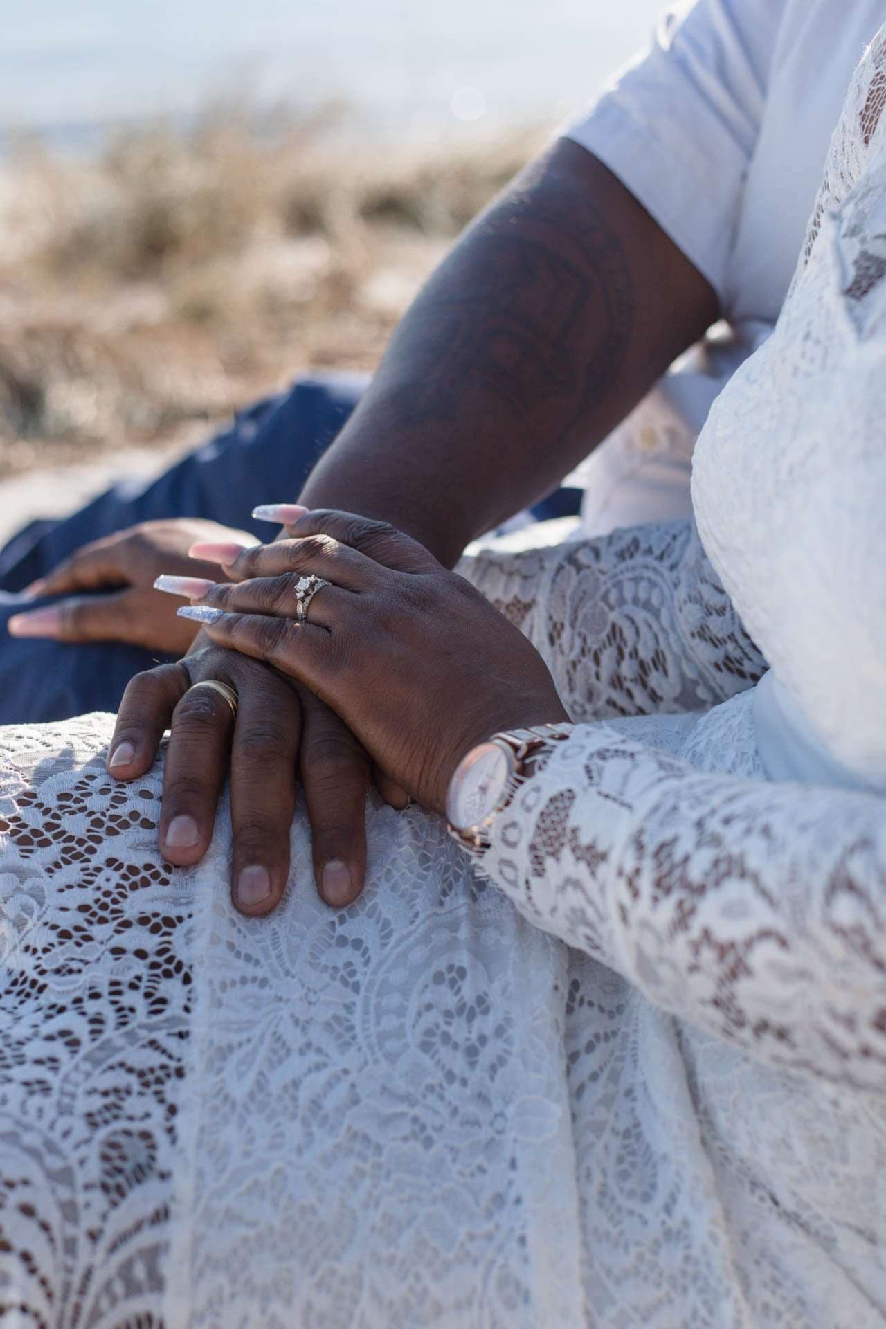 Close-up of a couple's hands with rings resting together on a white lace wedding dress outdoors