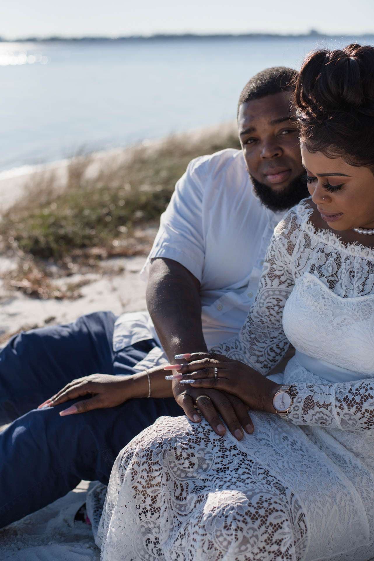 Couple sitting close on a sandy shore, her in a white lace dress with hands resting together, water behind them