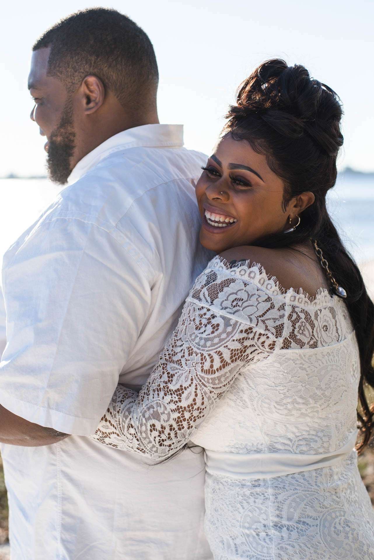 Smiling bride in an off-shoulder white lace dress hugging her partner in a white shirt beside the water