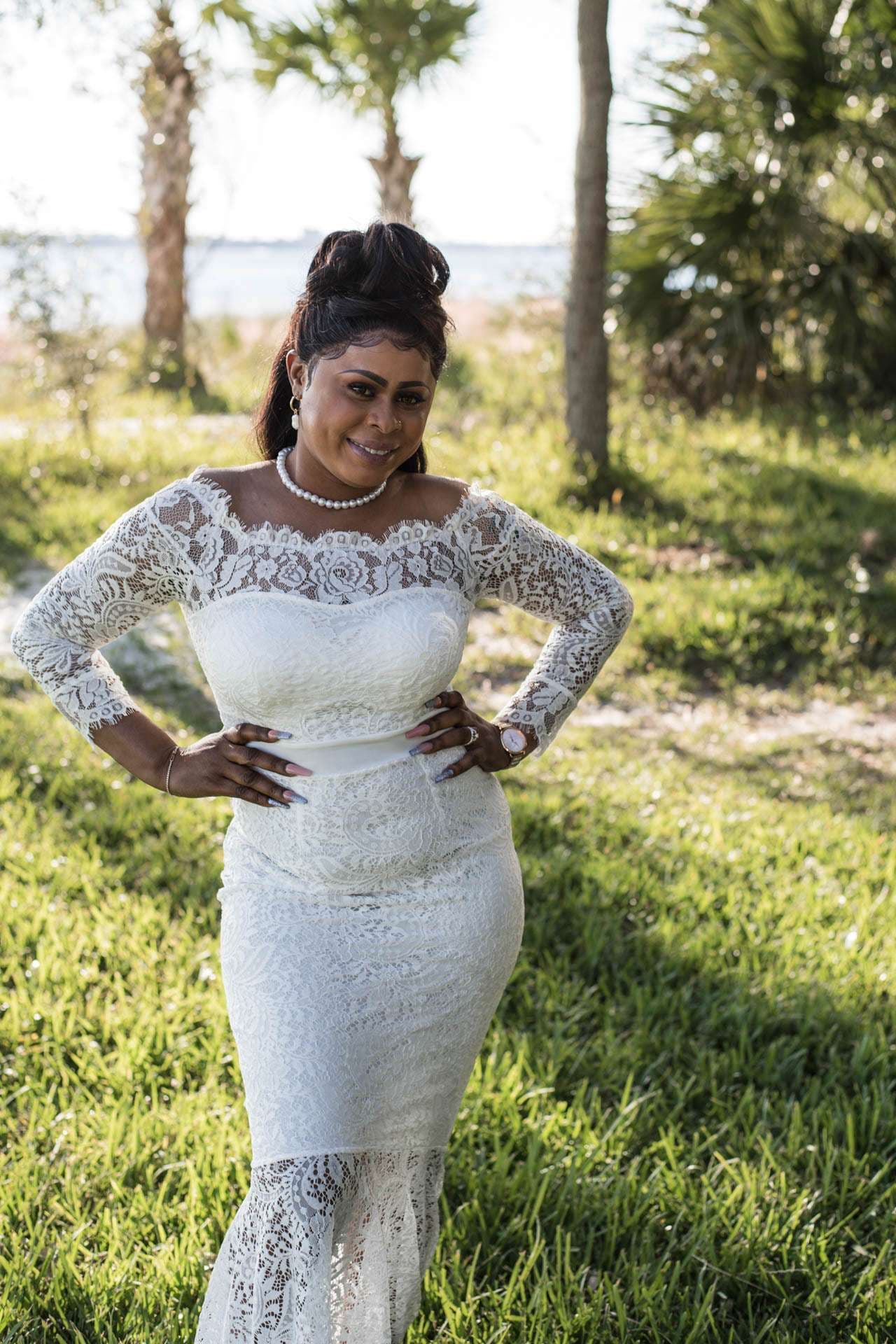 Smiling bride in a white lace off-the-shoulder gown and pearl necklace posing with hands on hips on a grassy lawn