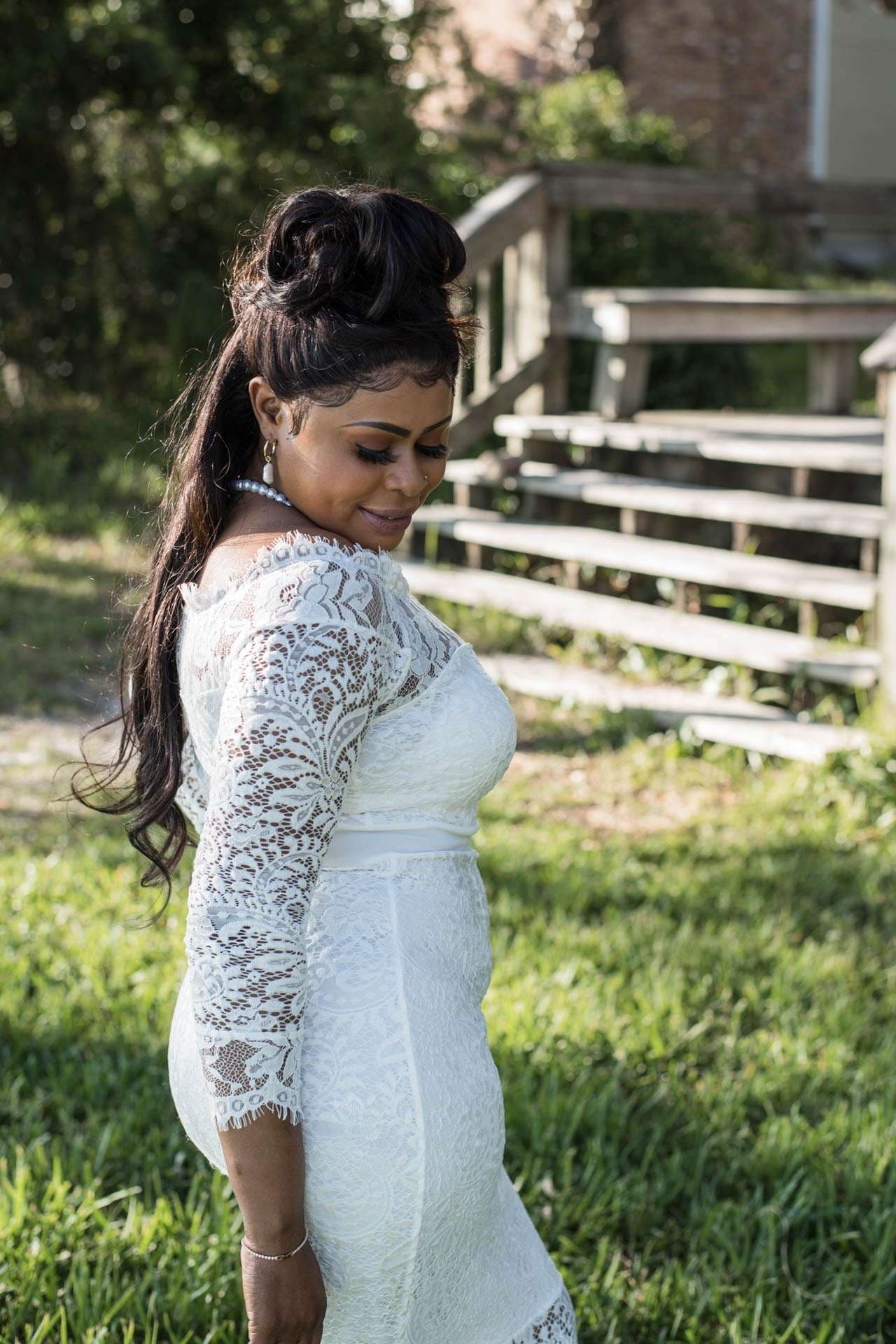 Woman in a white lace dress with a high bun and long ponytail glancing down on a grassy lawn near wooden steps