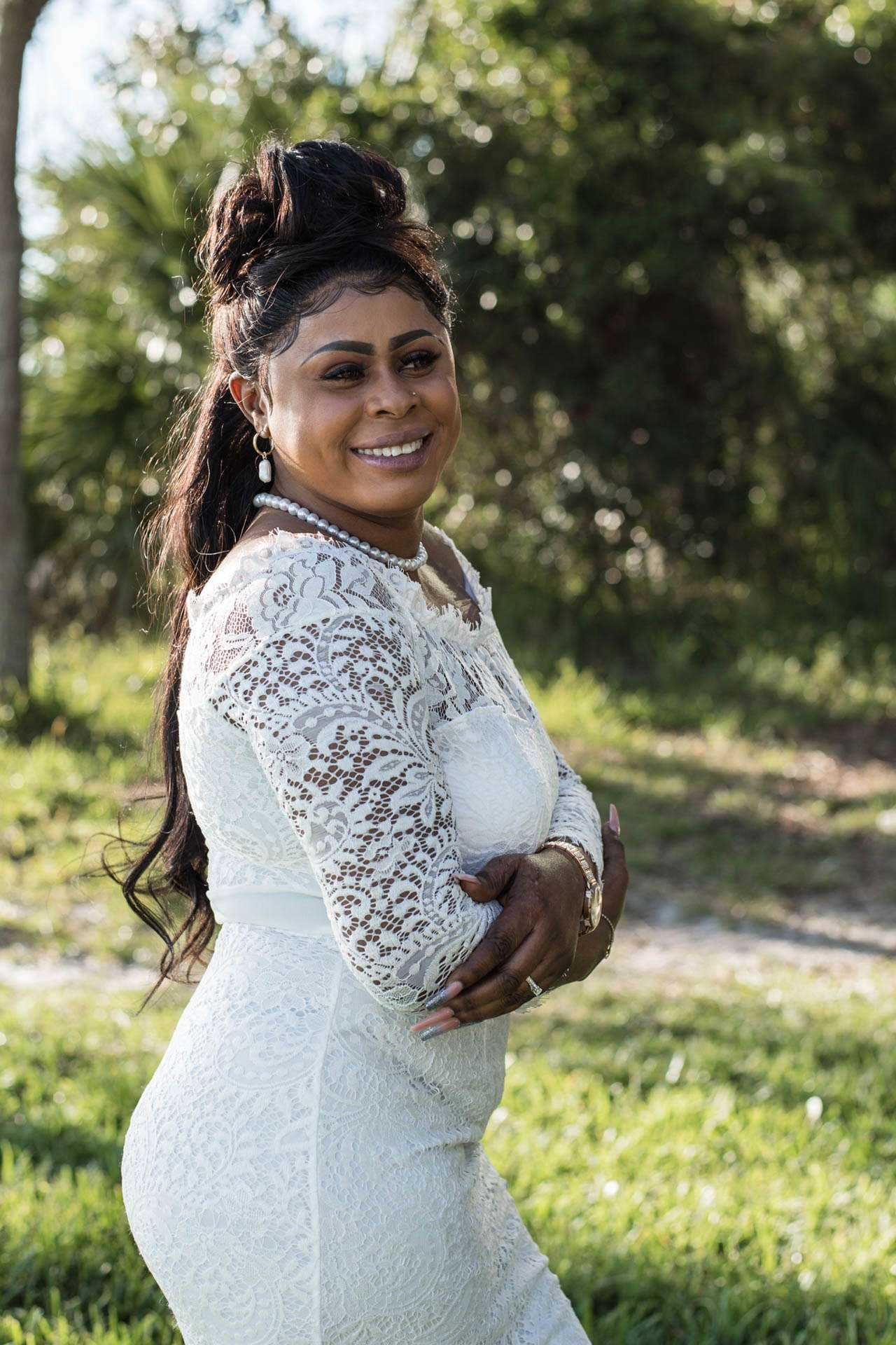 Smiling woman in a white lace long-sleeve dress and pearl necklace, arms crossed, standing on grass outdoors
