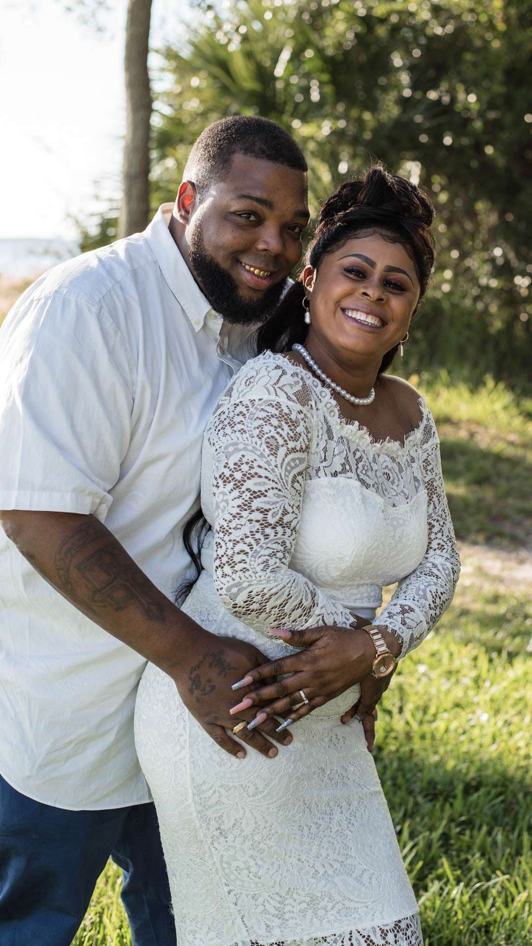 Smiling couple in white embracing outdoors on grass, her in a lace dress with pearl necklace, his hands on her waist
