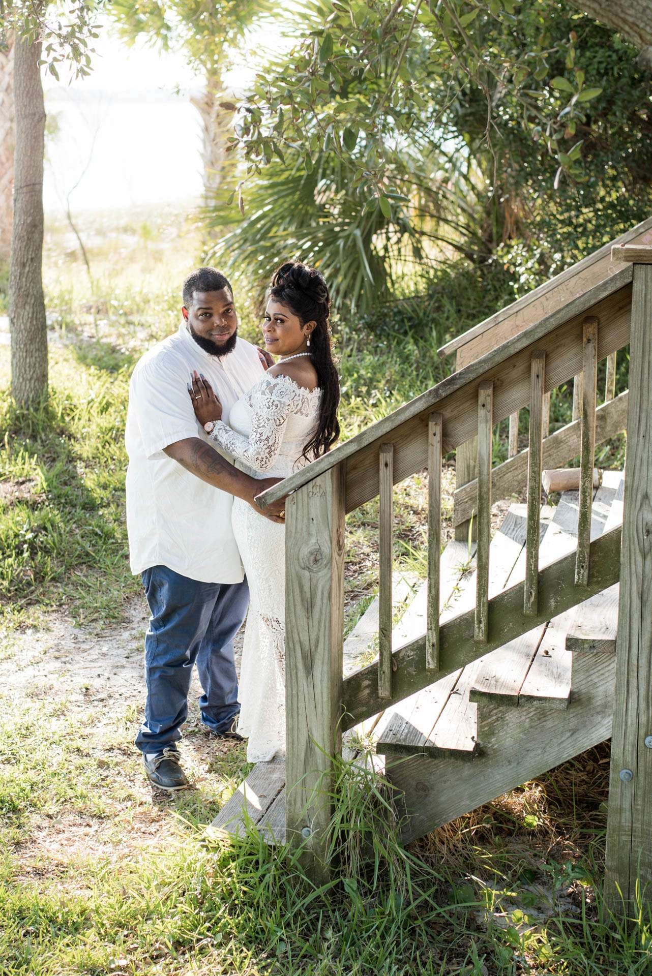 Couple embracing beside a wooden staircase outdoors, woman in a white lace gown, man in white shirt and jeans