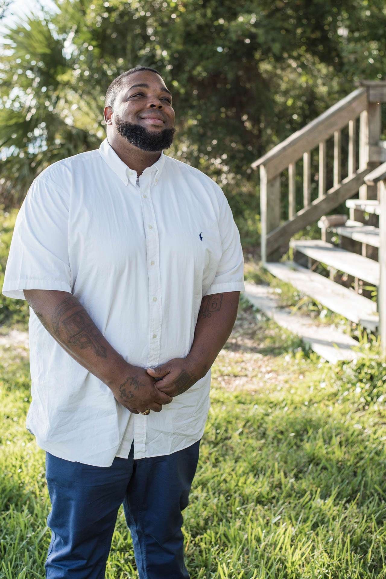 Smiling man in a white button-up shirt and blue pants standing with clasped hands on grass near wooden steps