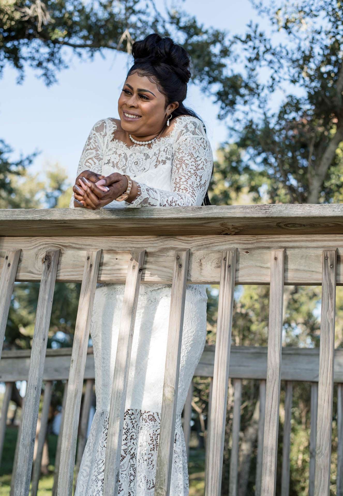 Smiling woman in a white lace gown leaning on a wooden railing beneath oak trees, gazing off to the side