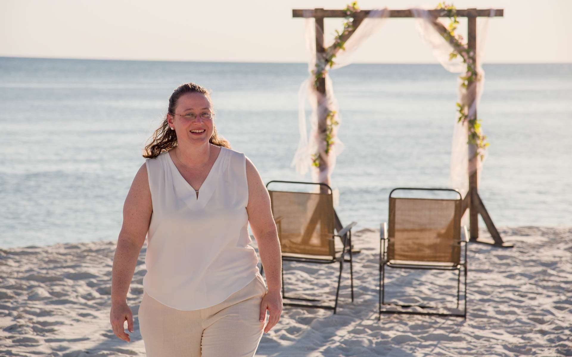 Smiling woman in a cream sleeveless top walks on the sand near a floral wedding arch and two beach chairs