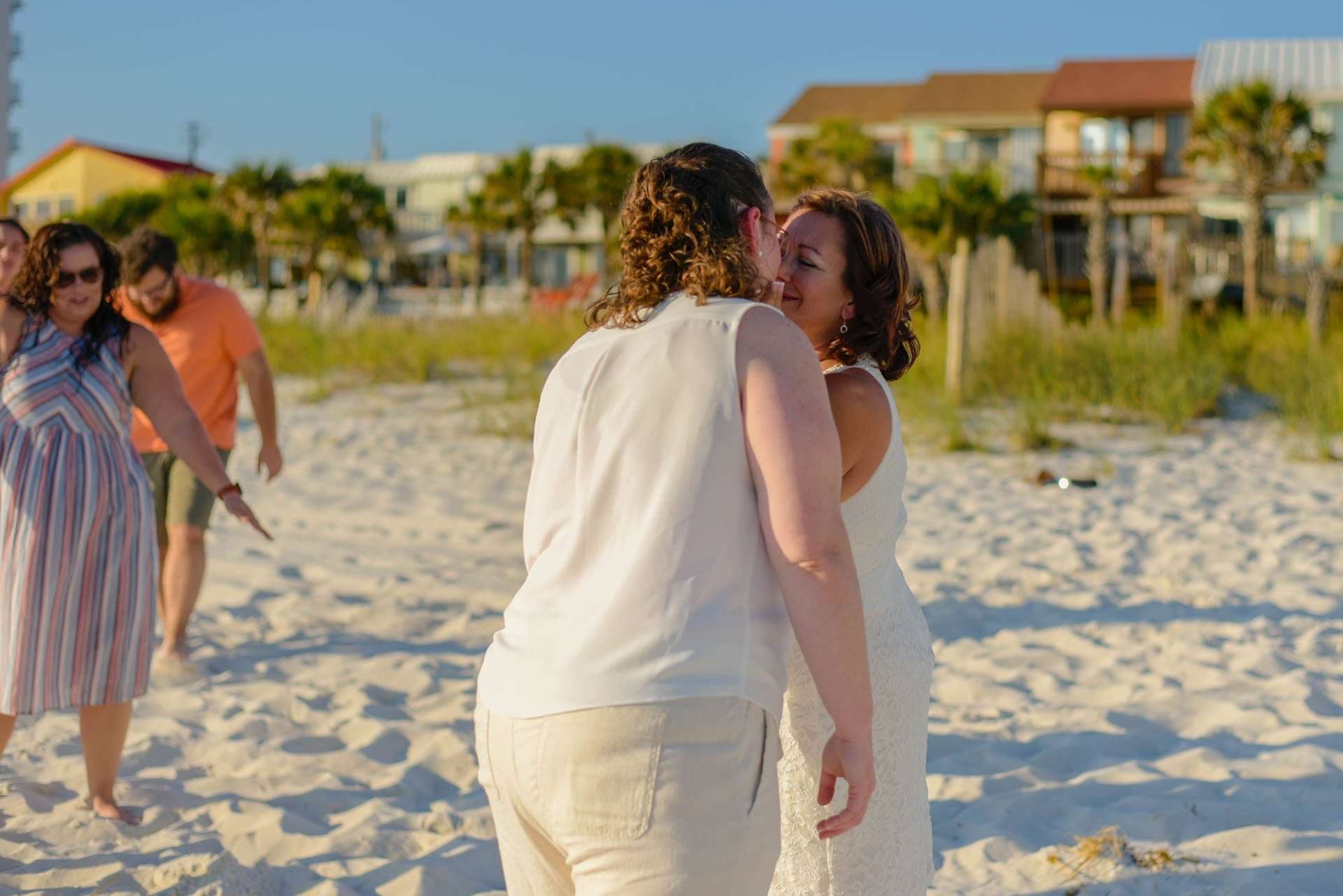 Two people in white outfits share a kiss on a sandy beach while guests walk behind them at golden hour