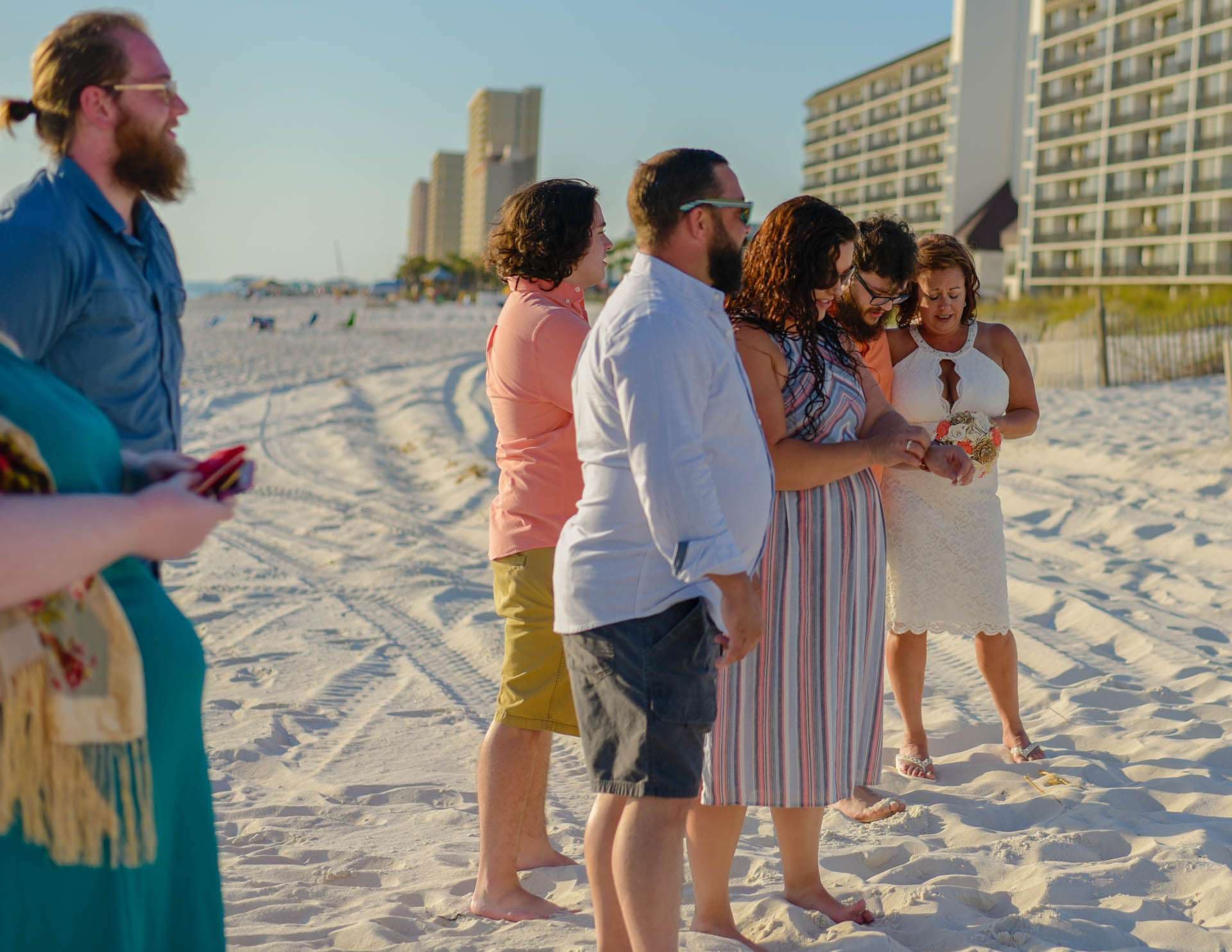 Barefoot guests gather around a woman in a white dress holding a bouquet at a beach ceremony