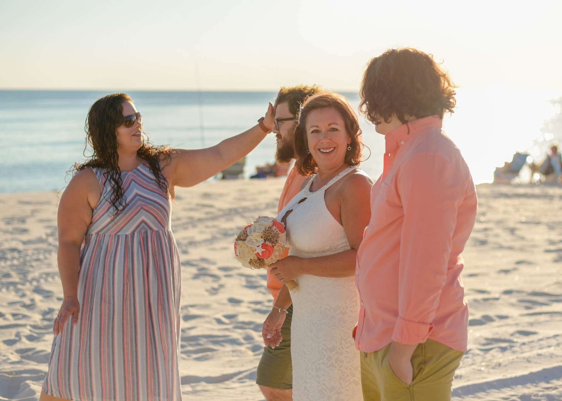 Smiling woman in white dress holding a bouquet on the beach as a woman in sunglasses reaches out beside her