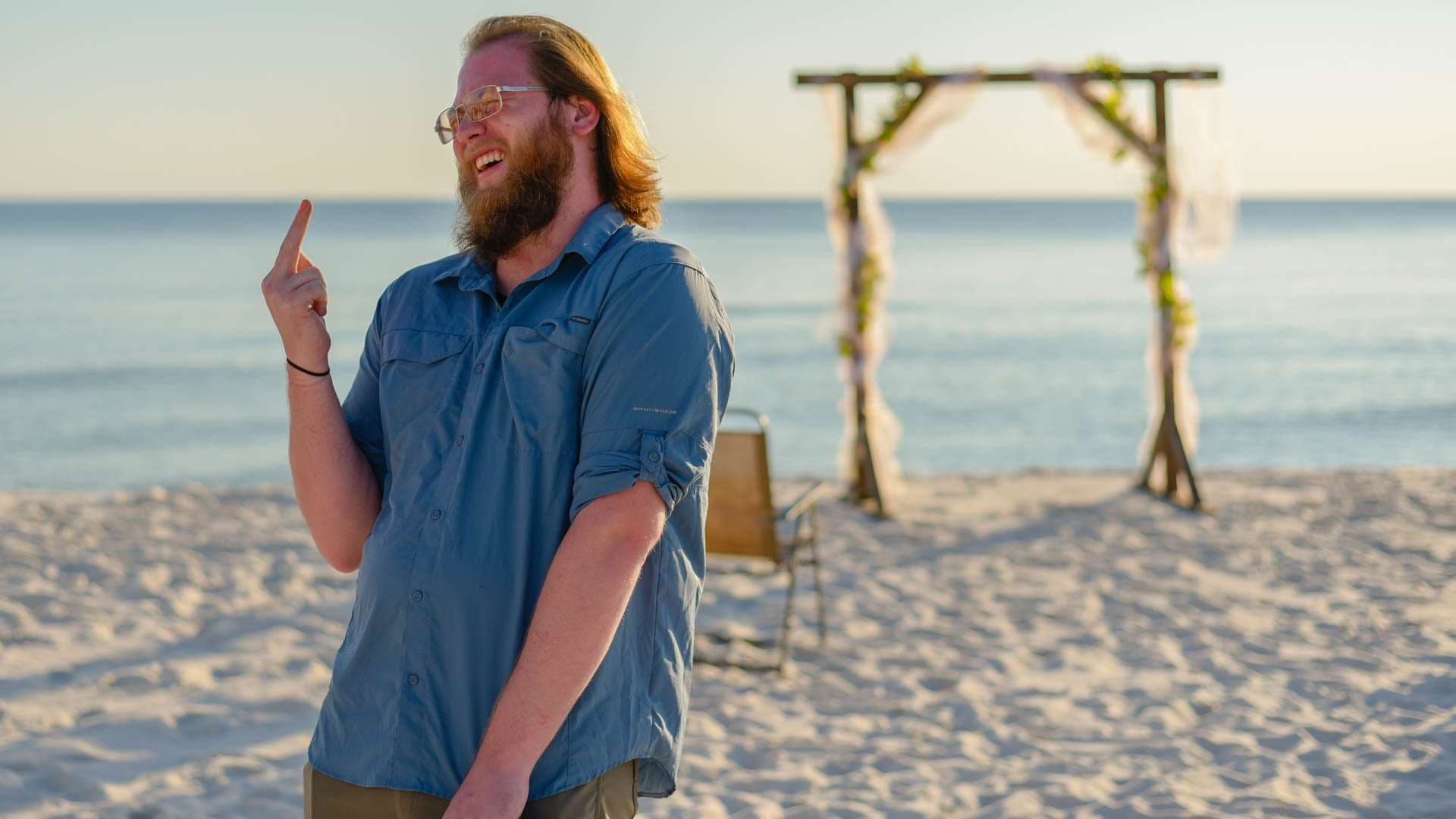 Bearded man in glasses and a blue button-up shirt laughing on the beach near a floral wedding arch
