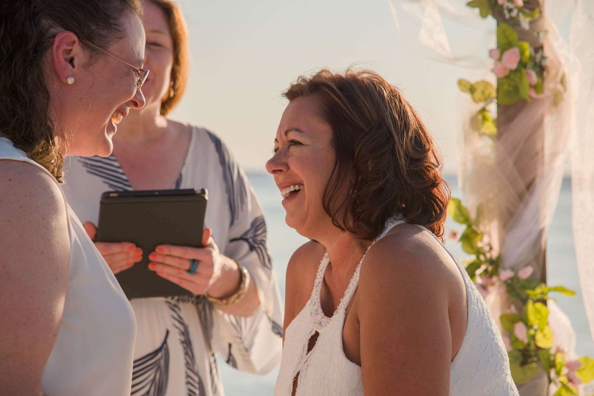 Two brides laughing during a beach ceremony as an officiant holds a tablet beside a floral arch by the water