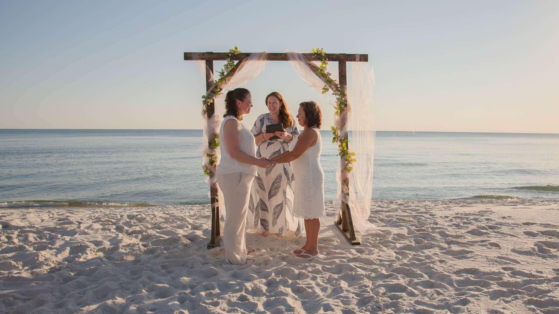 Two brides in white holding hands under a flowered wooden arch on the beach while an officiant stands between them