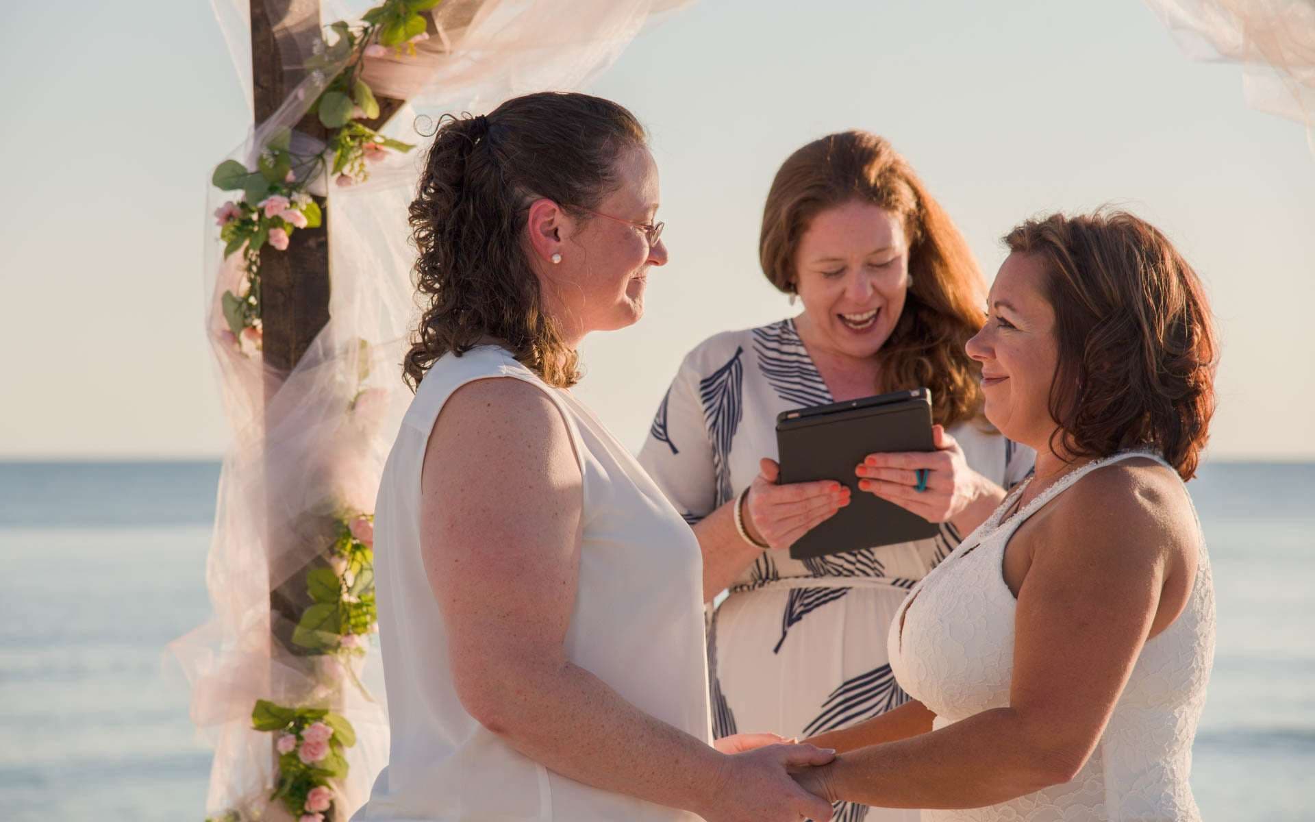Two brides in white holding hands during their beachside ceremony while a smiling officiant reads from a tablet
