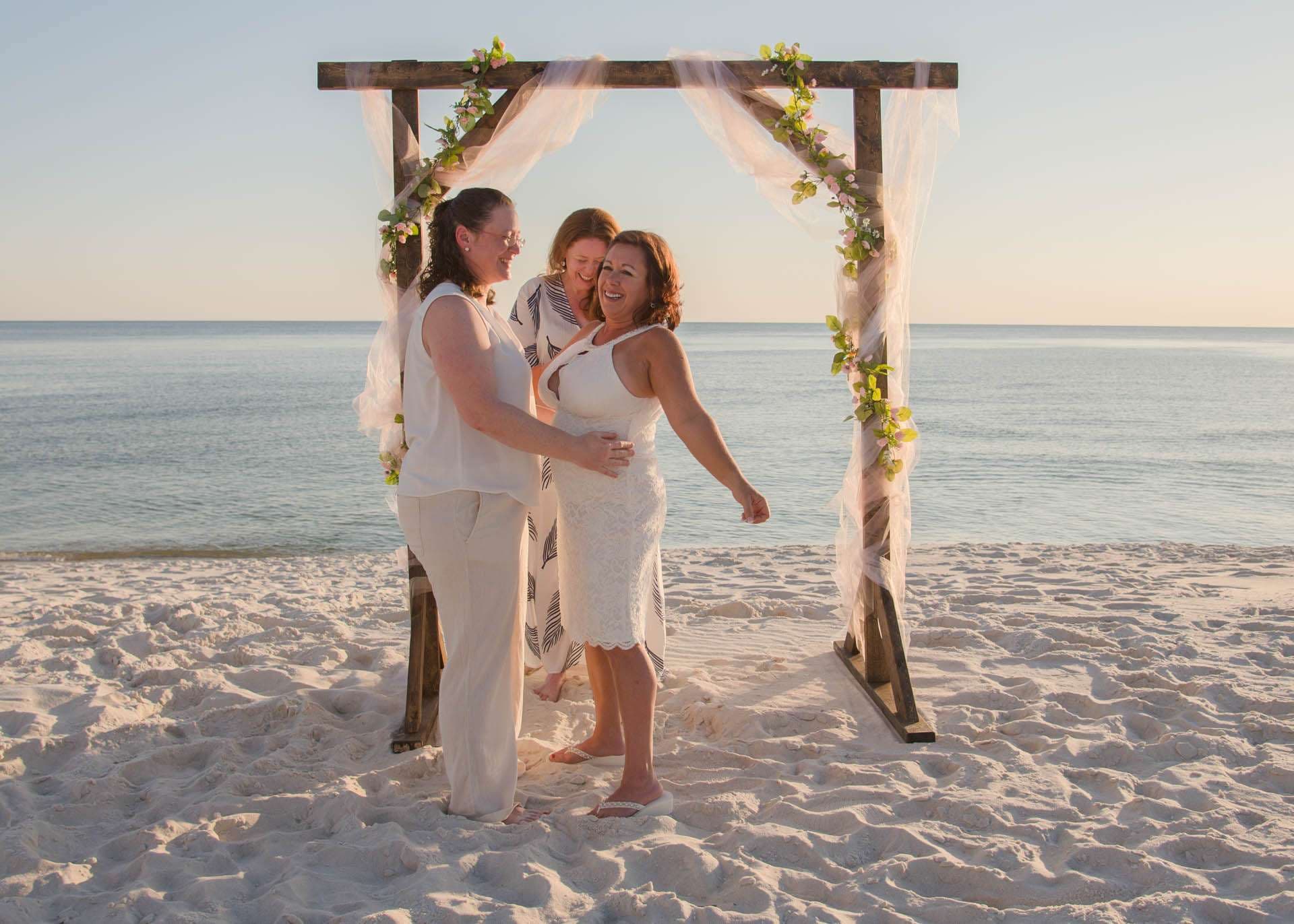 Two women embracing under a wooden beach arch with tulle and greenery while an officiant smiles behind them