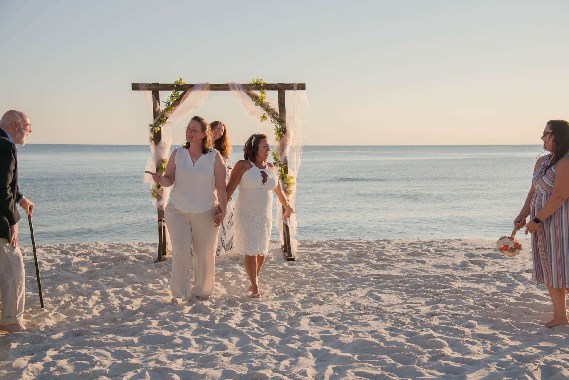 Two brides holding hands and walking from a floral wooden arch on the sand as guests watch