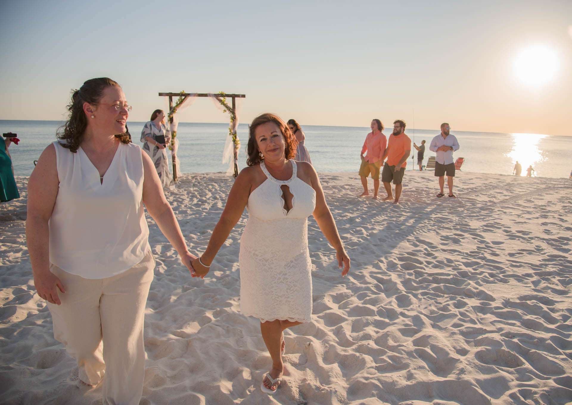 Two women in white holding hands and walking across the sand at sunset, with a floral wedding arch behind them