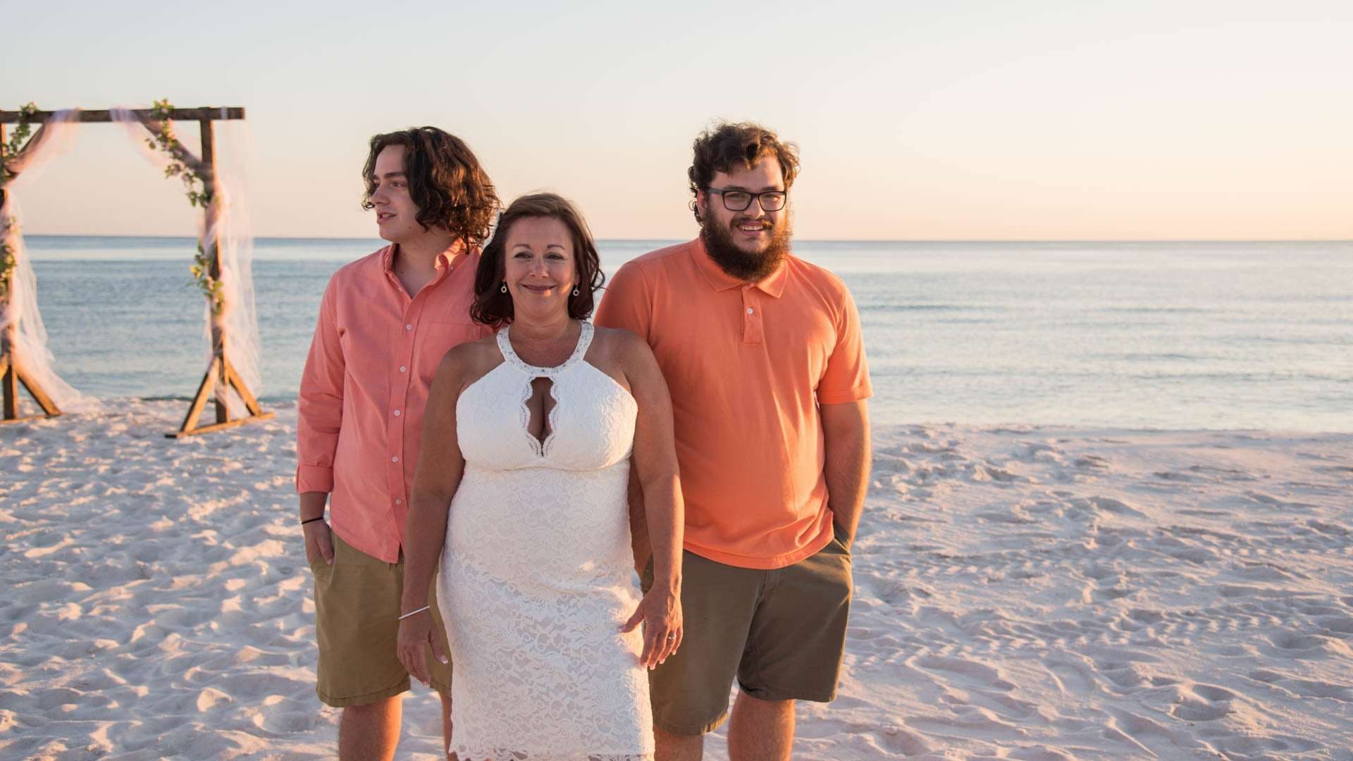 Woman in a white lace dress standing between two young men in orange shirts on the beach near a wedding arch