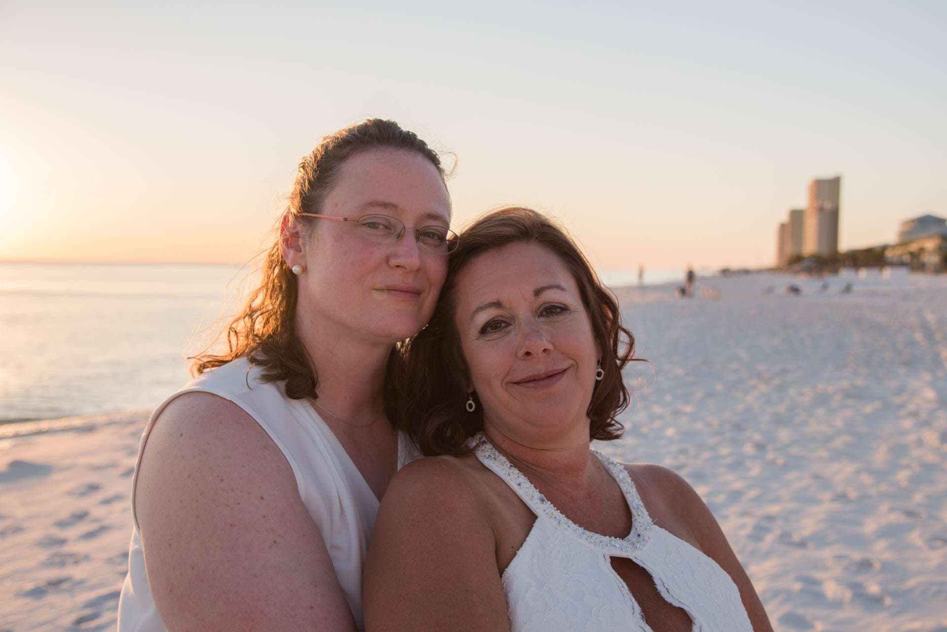 Two women in white tops embracing and smiling on a sandy beach at sunset