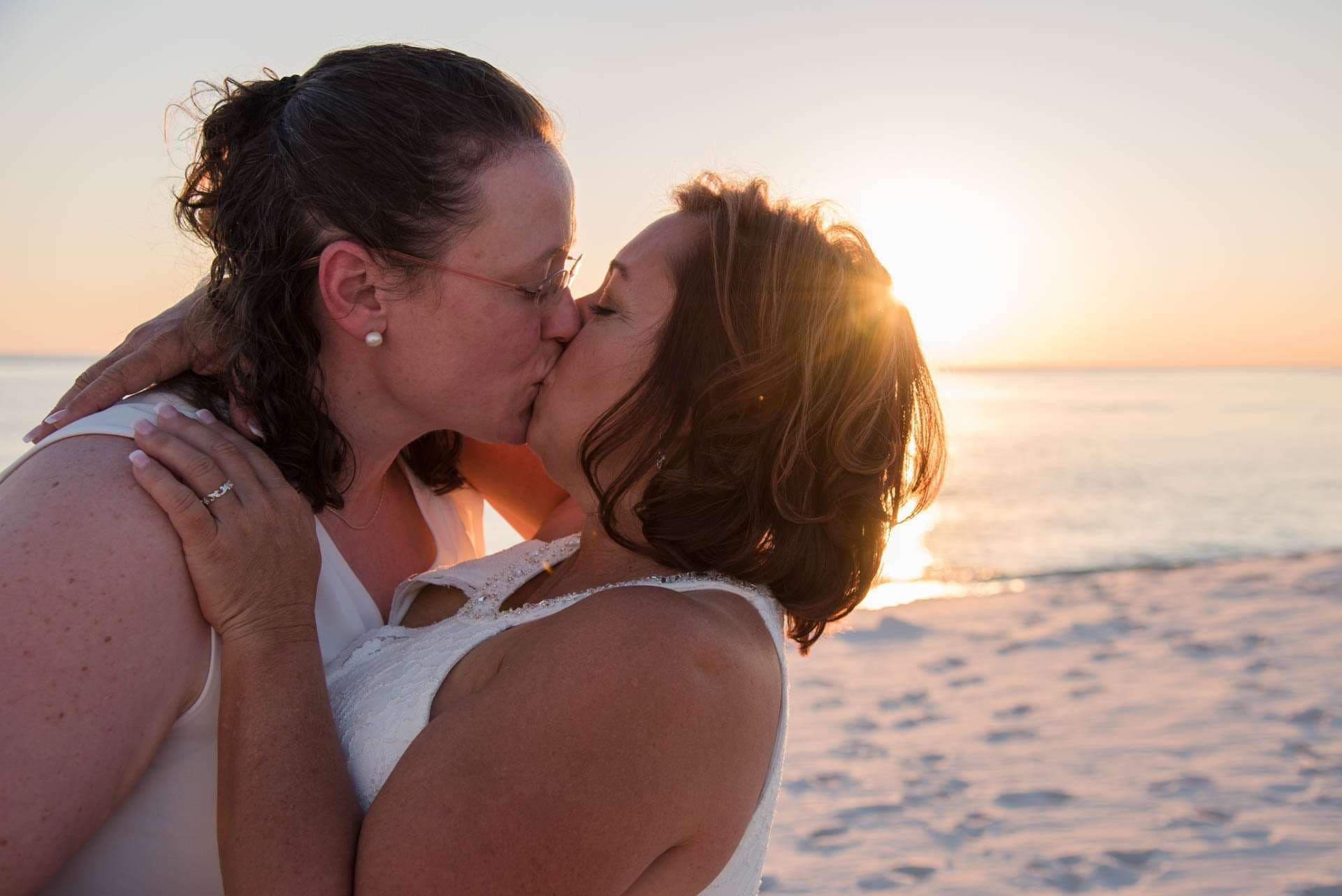 Couple in white kissing on the beach at sunset, sun glowing over the water behind them