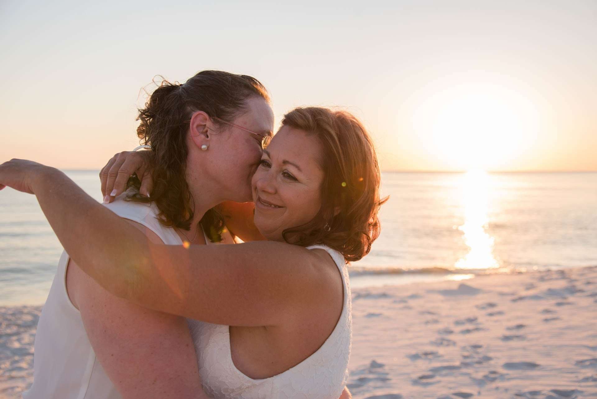 Two women in white embracing, one kissing the other's cheek, on a sandy beach at sunset