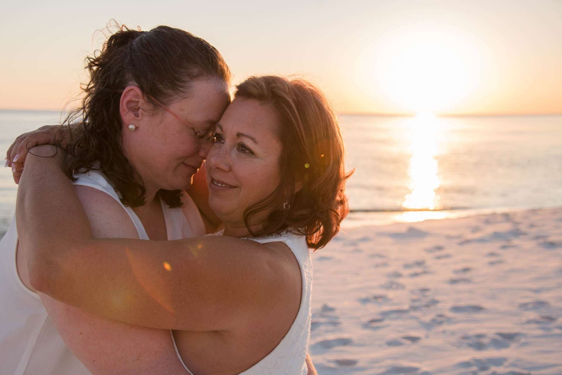 Two women in white embracing forehead to forehead on the beach as the sun sets over the water