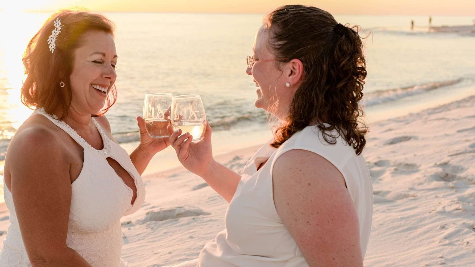 Two brides in white laughing and toasting with wine glasses on the beach at sunset