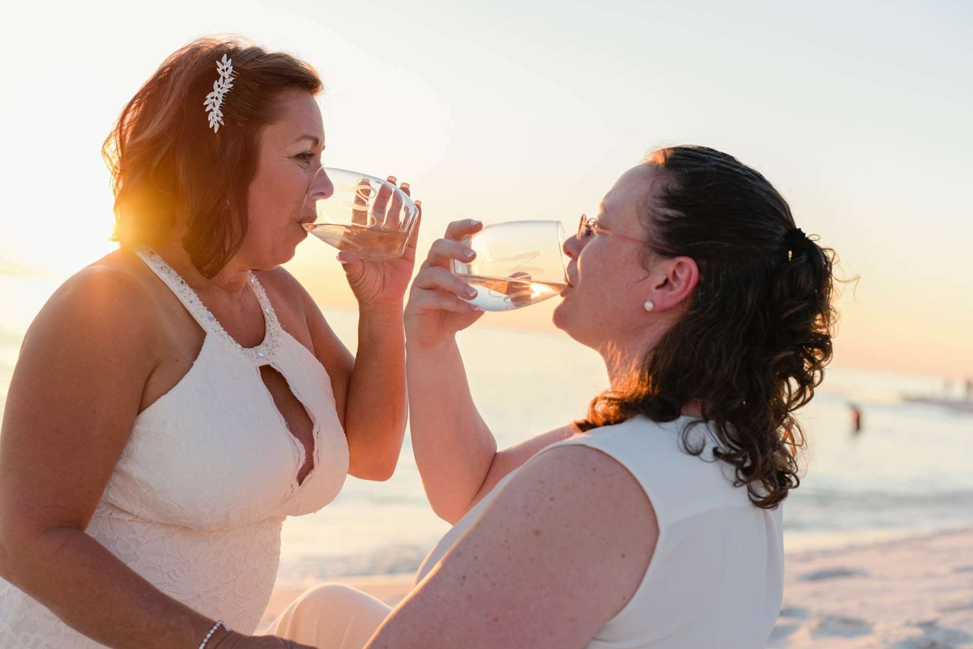 Two women in white drinking wine from glasses on the beach at sunset