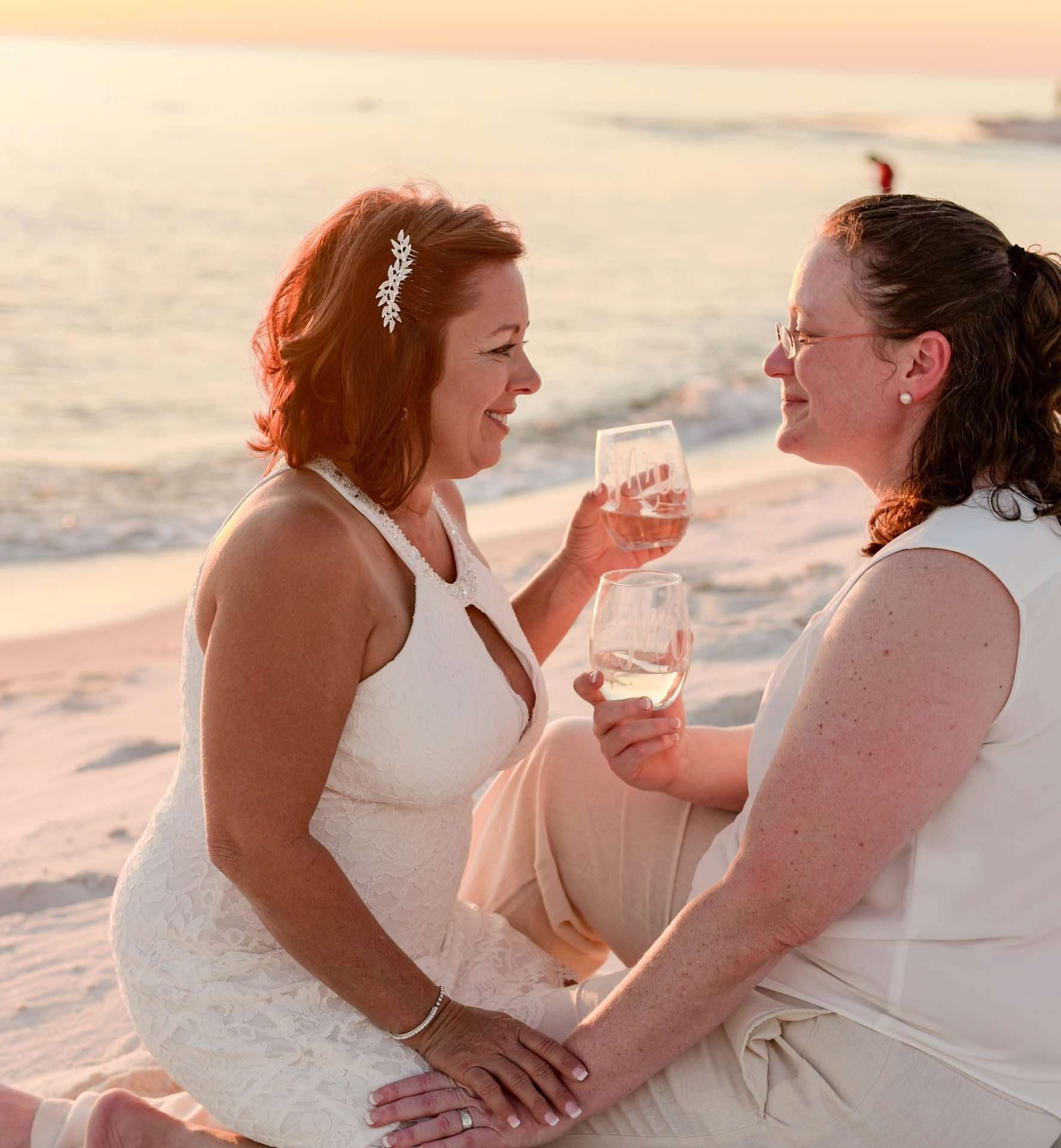 Two women in white smiling at each other and holding wine glasses on the beach at sunset