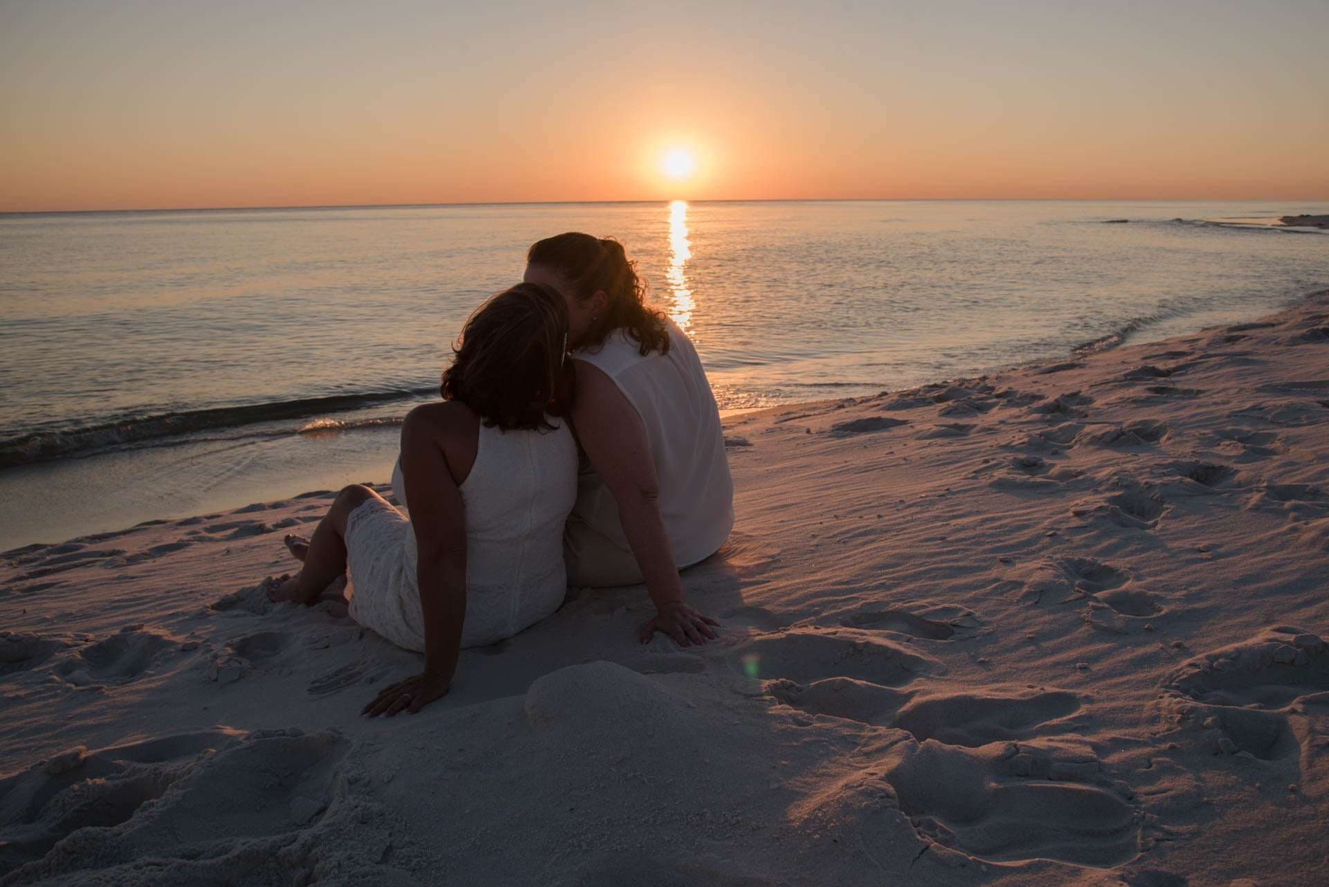 Couple in white sitting close on the sand, one kissing the other's head, watching the sun set over the water