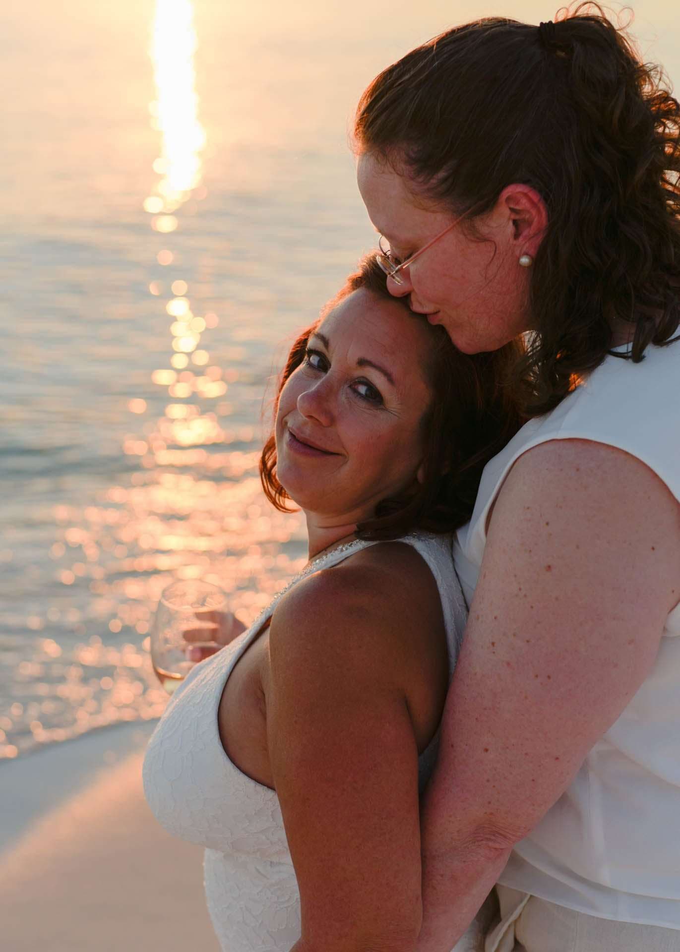 Two women in white embracing on the beach at sunset, one kissing the other's forehead while she holds a wine glass