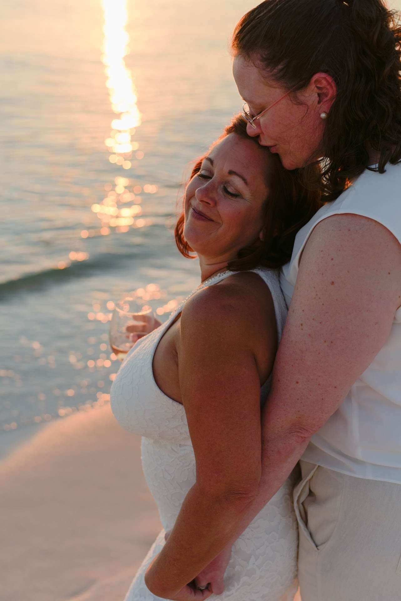 Two brides embracing on the beach at sunset, one in a white lace dress kissing the other's forehead